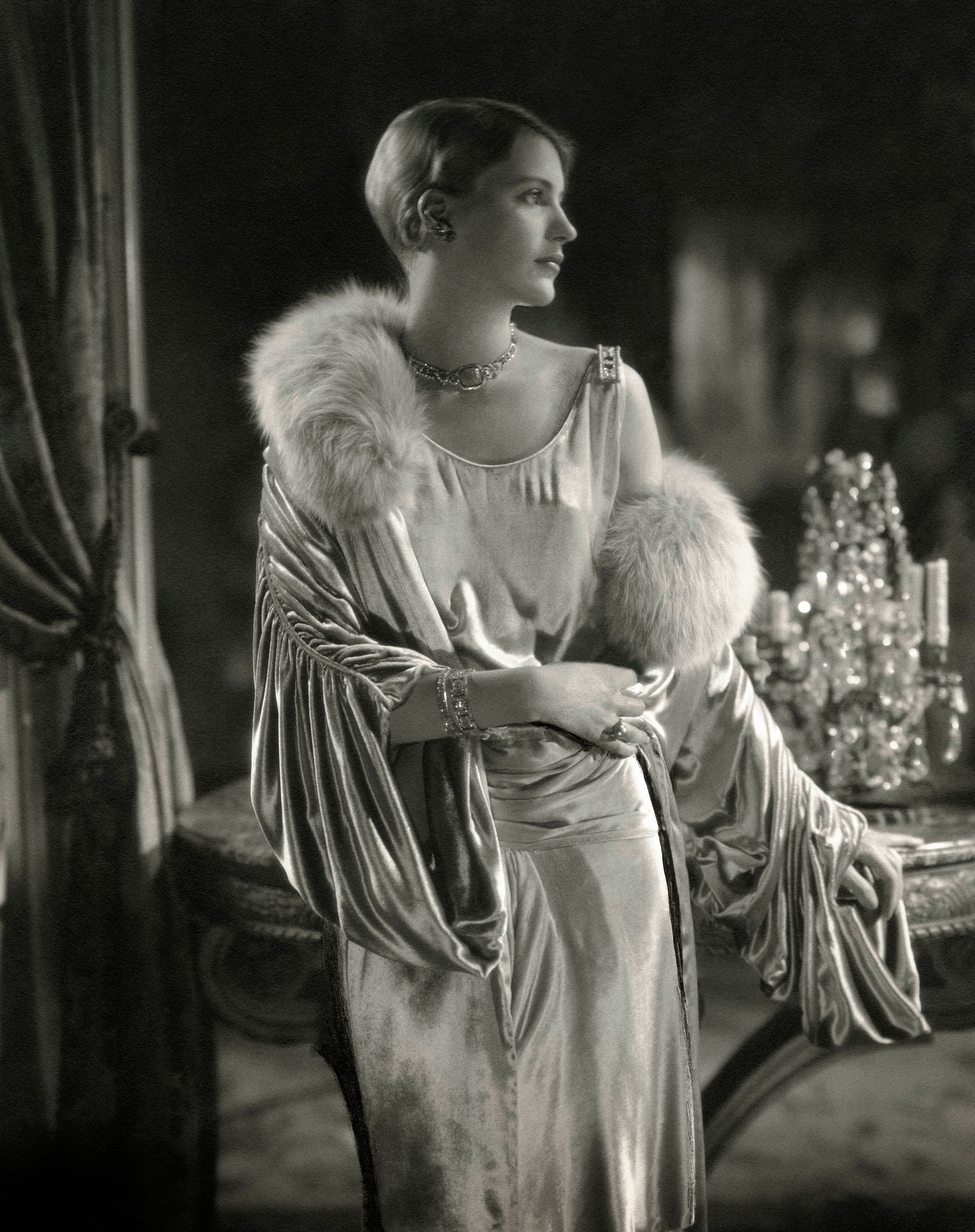 A white woman dressed in a fur stole and velvet gown stands in front of a gilt table.
