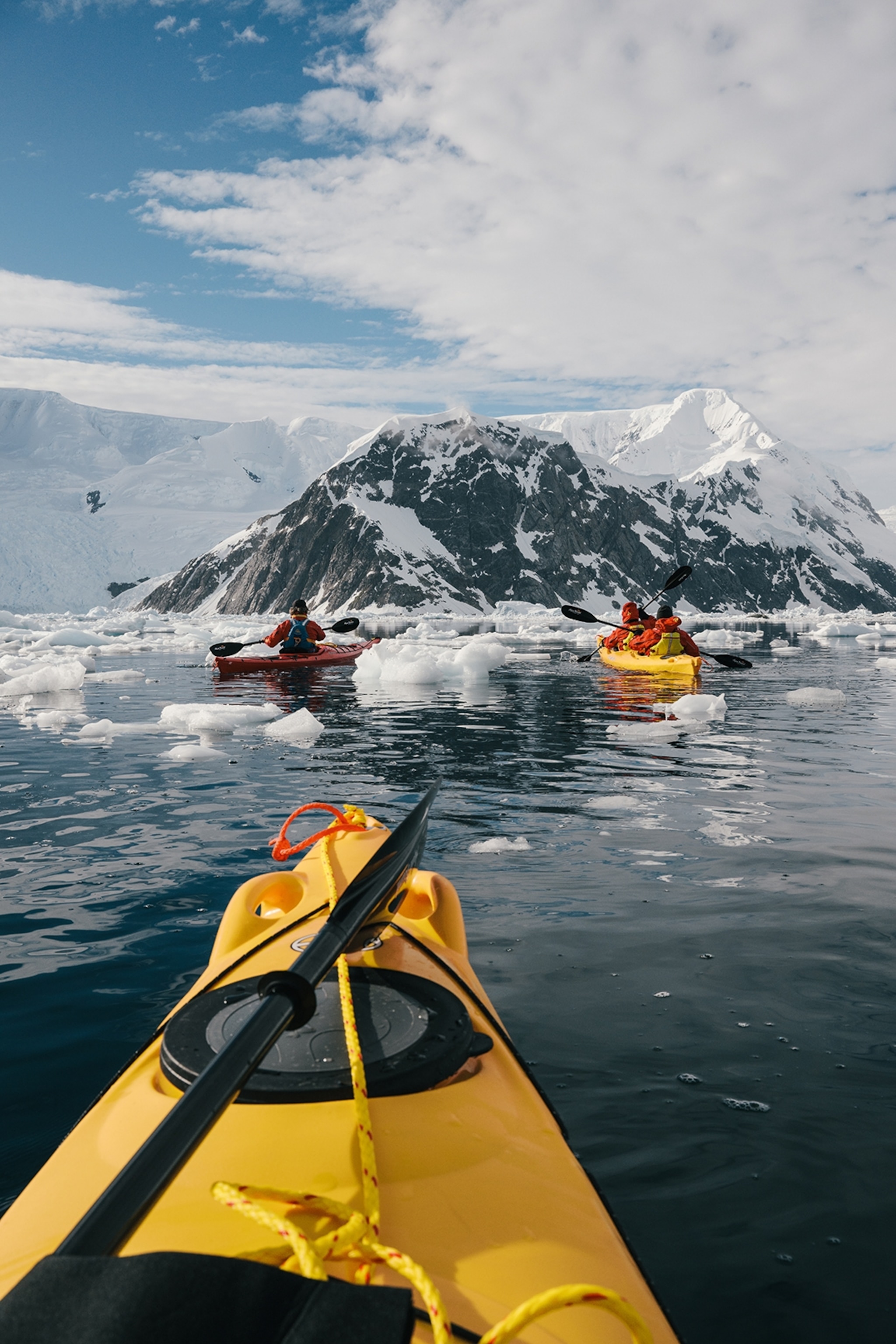 A view from a kayak in Antarctica. The kayak is yellow, and chunks of ice float in the water. in front is a snow-topped mountain.