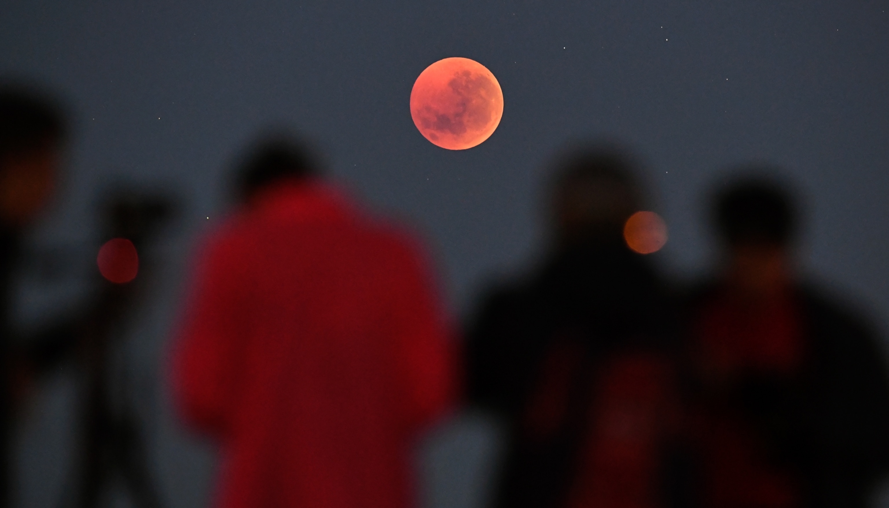 A group of people are seen out of focus in the foreground as they observe a bright, red, full moon in the sky above.