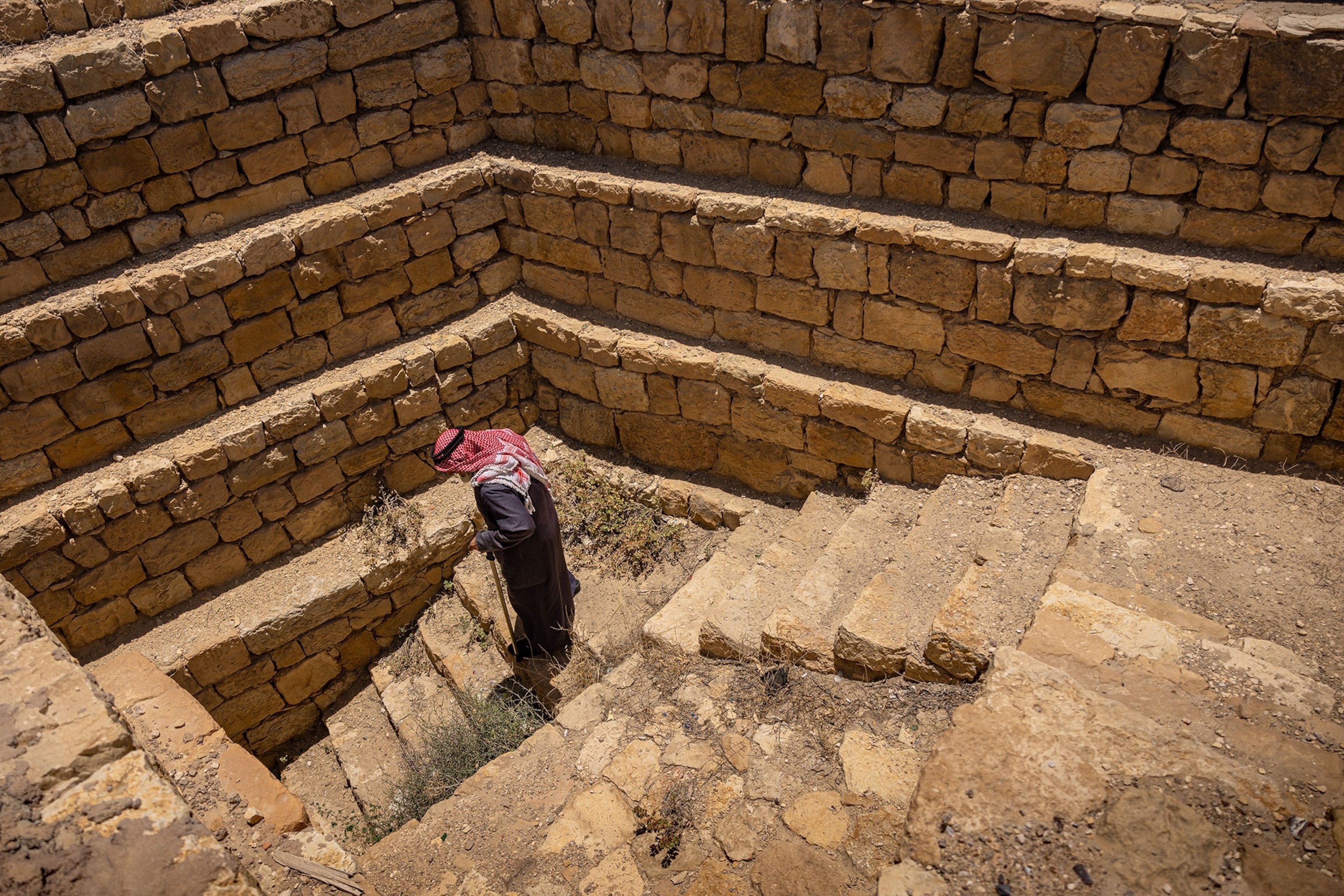 A man, with a red headdress, grey robe and cane, walks down stars surrounded by layers of wall stones.