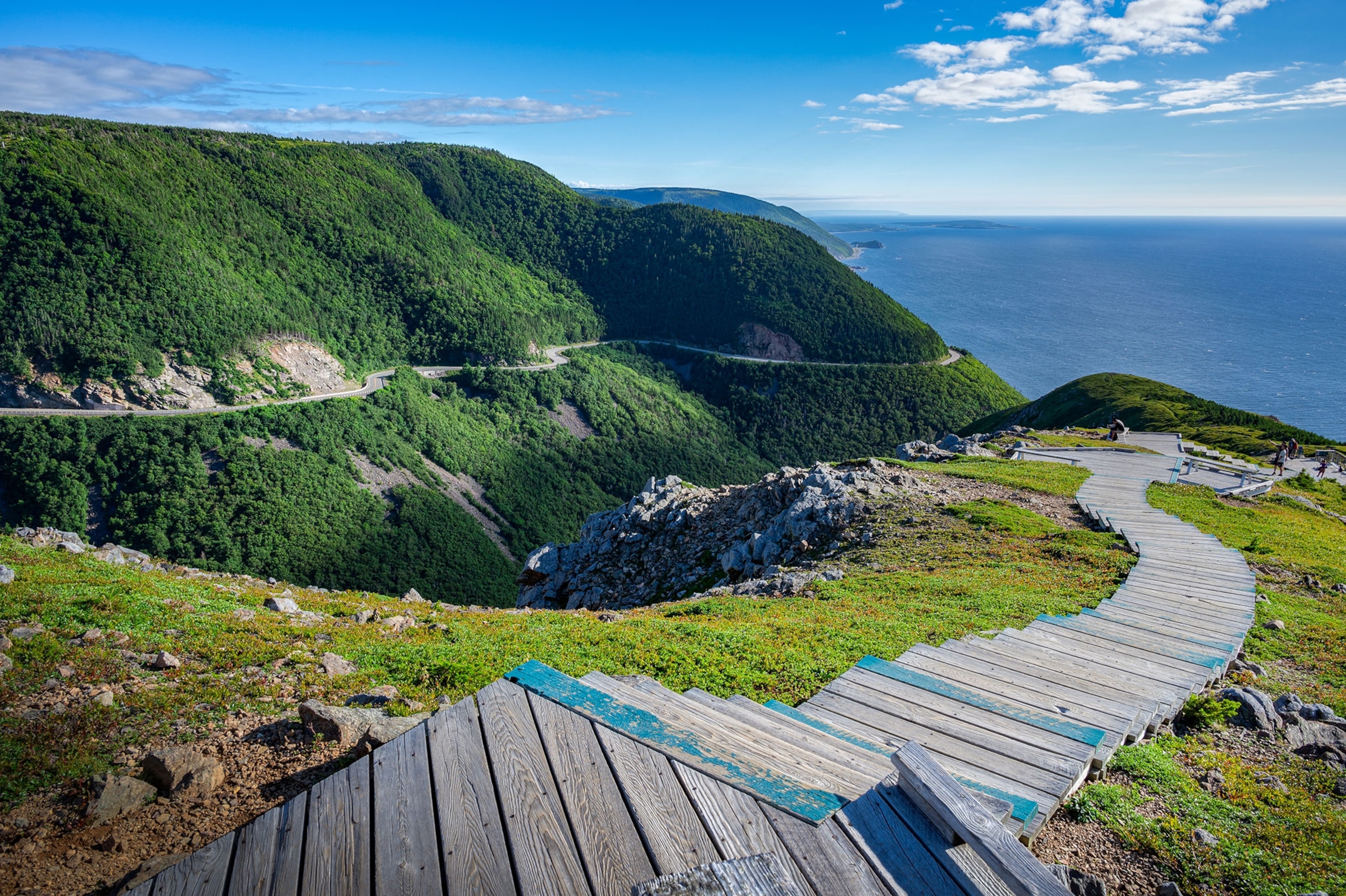 A wooden trail over rocky, green hills, leads down to a blue ocean. A winding road and tall, tree covered hills are in the background.