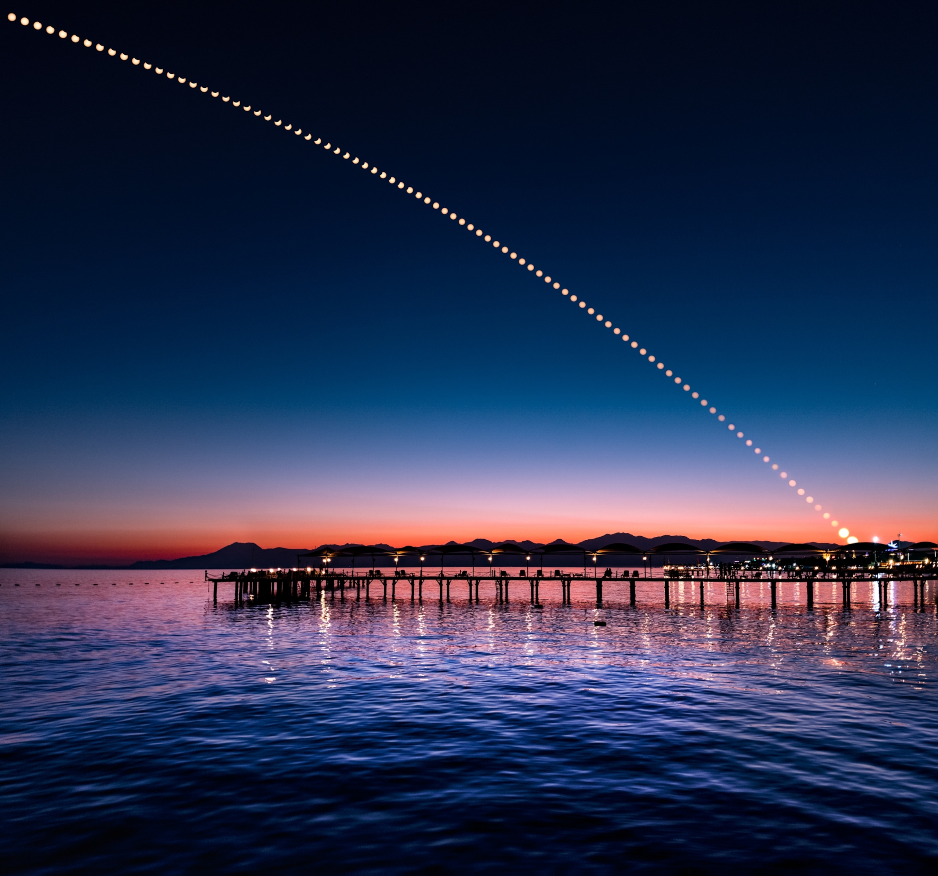 A silhouetted dock at sunset with mountain in the background. The sky transitions from deep blue at the top, to fiery reds behind the mountains on the horizon. Across the sky, a sequence of the Sun is seen as it is partially eclipsed by the Moon.