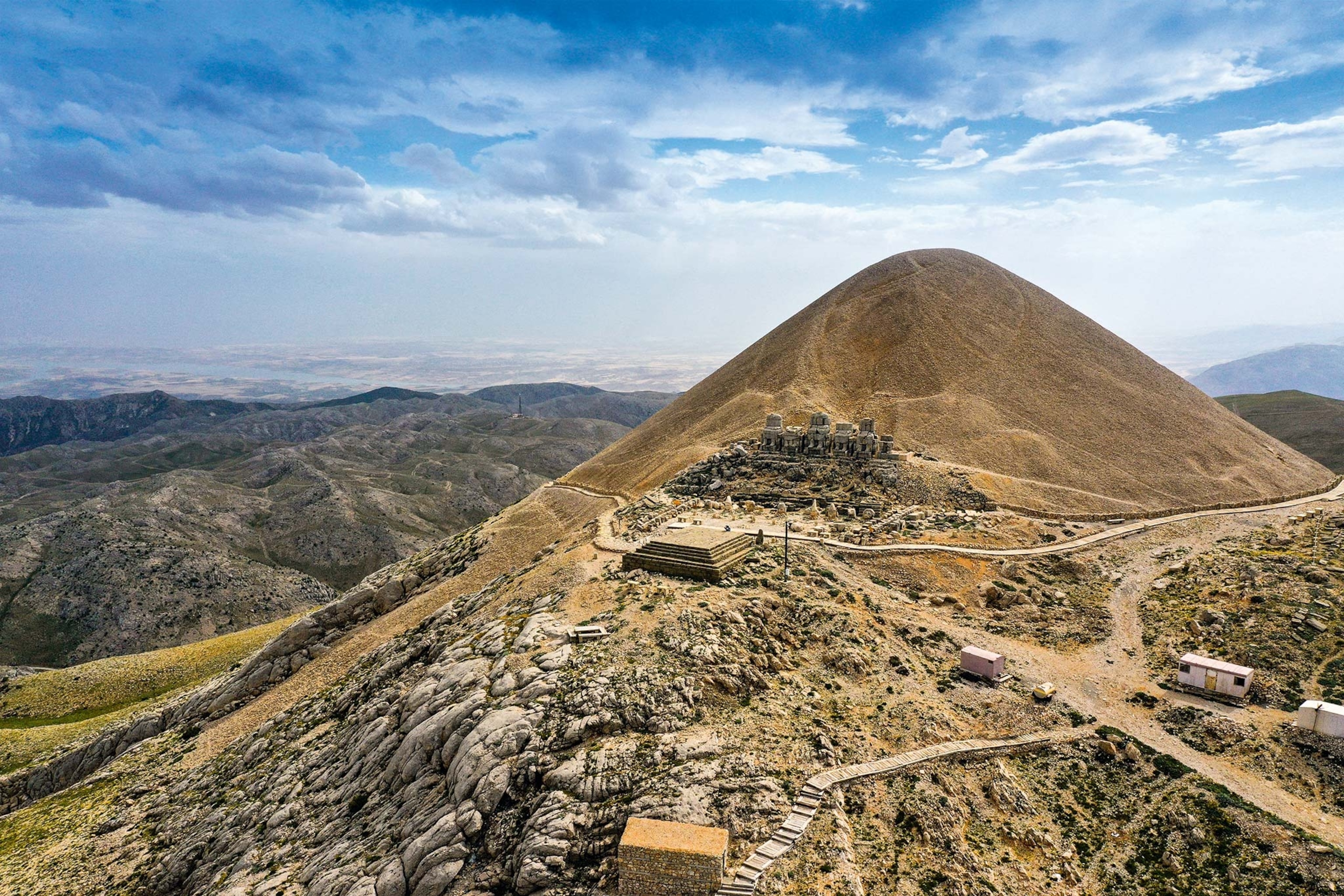 Behind terraces and statues lies a large mound, with trailers set up for excavation and a blue sky