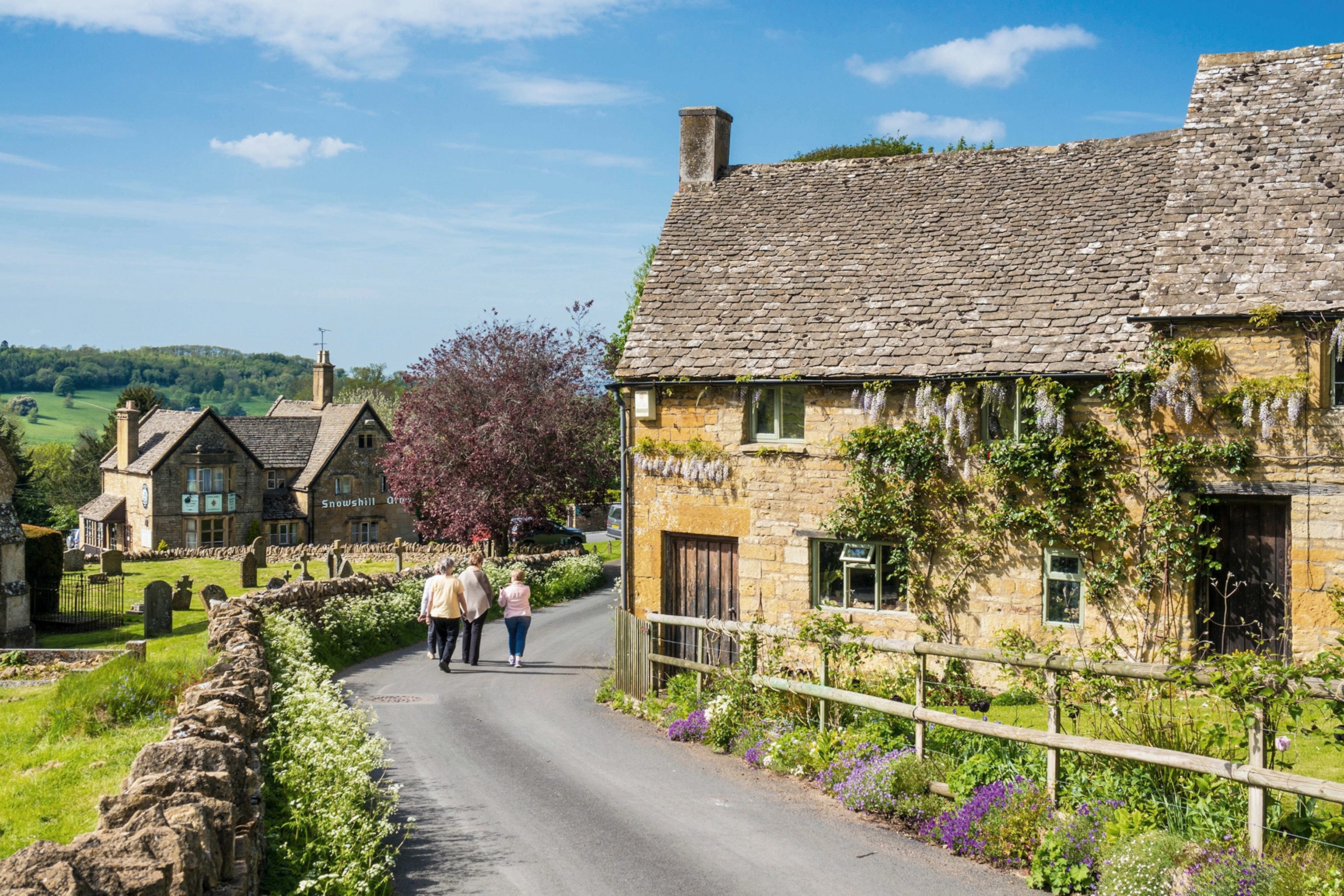 A group of people strolling down the snaking country road past ancient stone cottages and lush greenery.