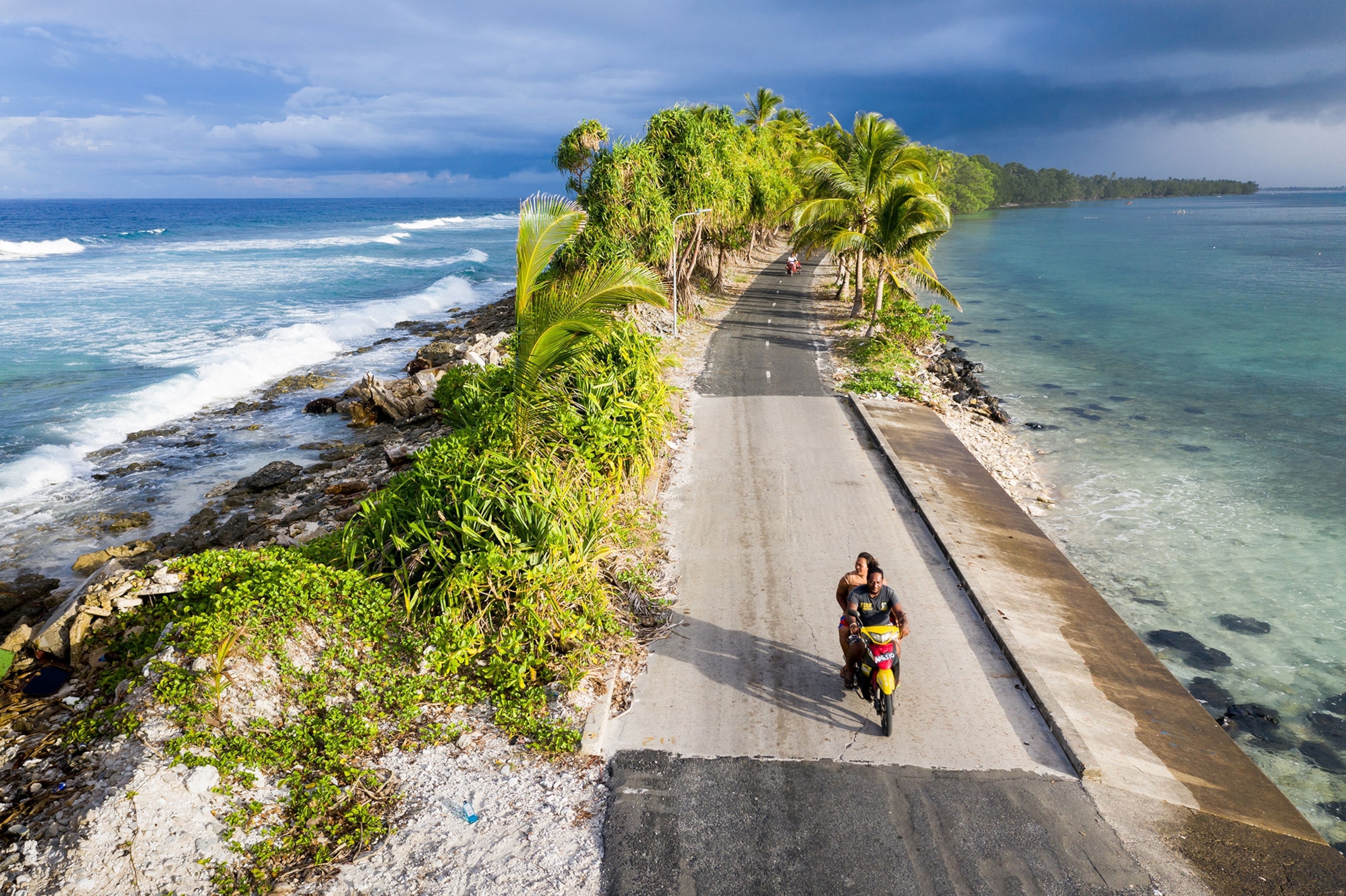 Two people ride on a motorbike down a narrow road surrounded by blue water; on the left is the Pacific ocean and the right, a lagoon. This road is lined with palm trees and is the narrowest point of Fongafale island in Tuvalu.