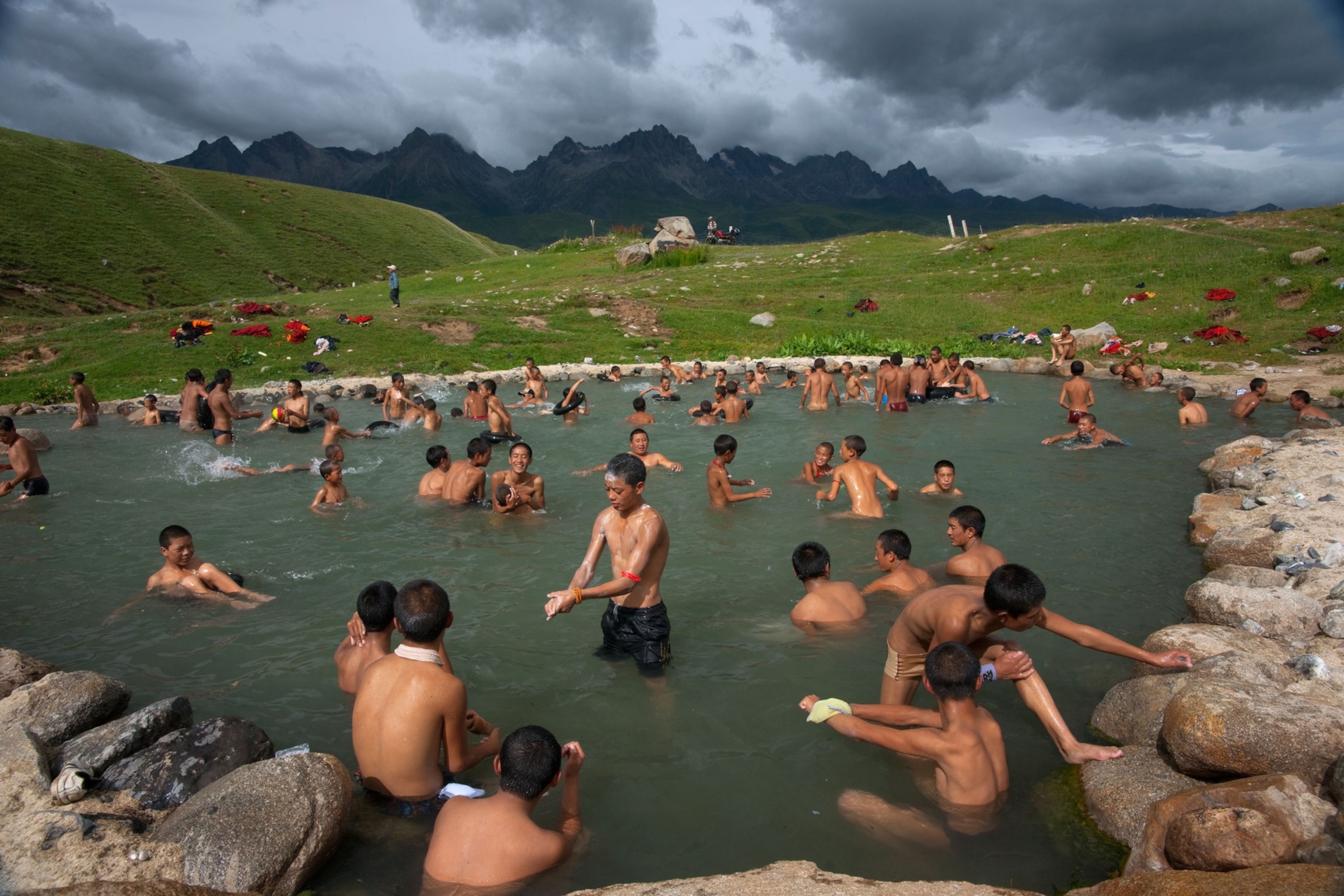 Several dozen boys and young men wearing black shorts bath in a pool of water surrounded by rock. In the distance is a green grassy knoll which extends into black mountains and grey clouds.