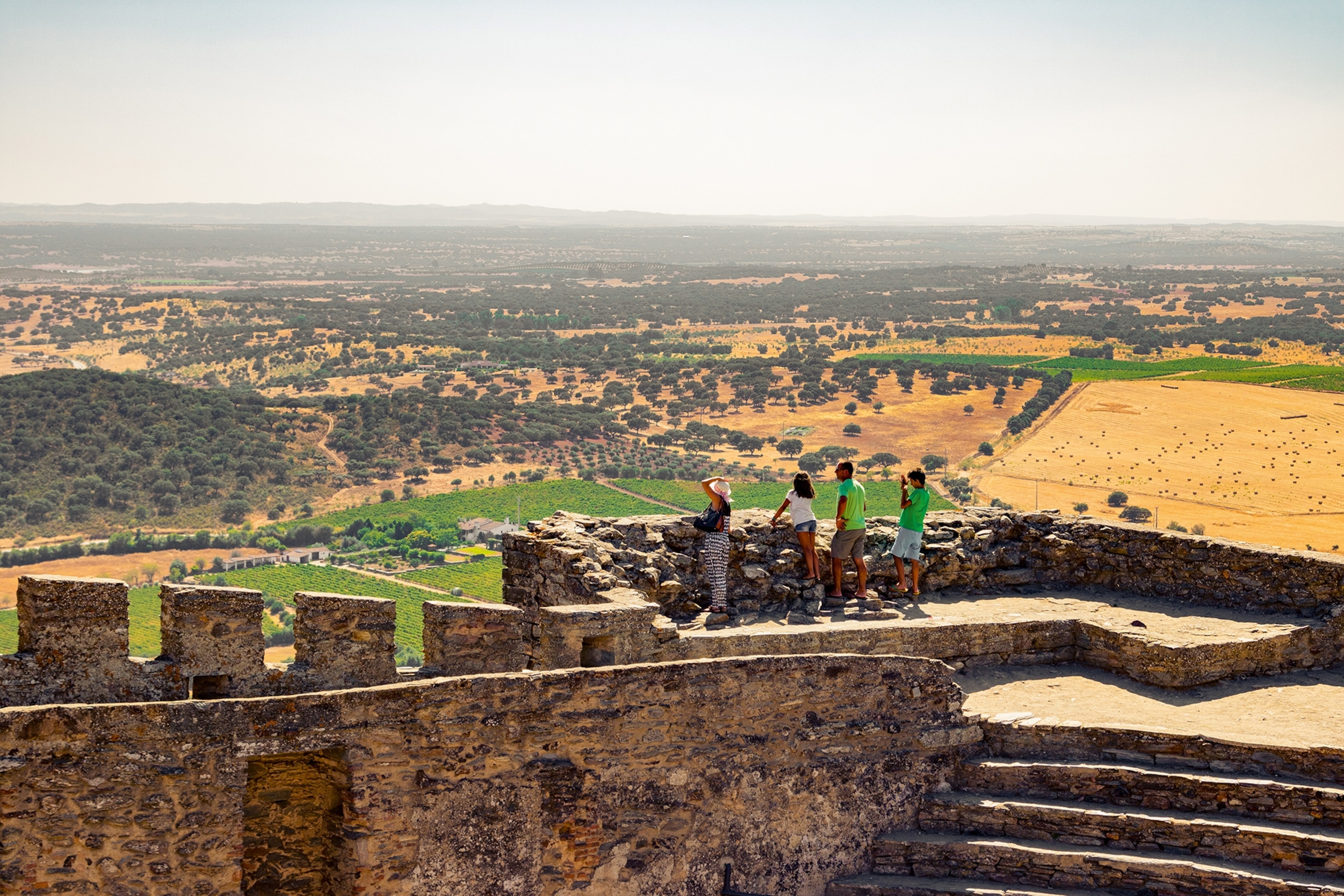 A wide view onto a walkable castle stone wall with a family looking at the view on the fields and bushy landscapes.