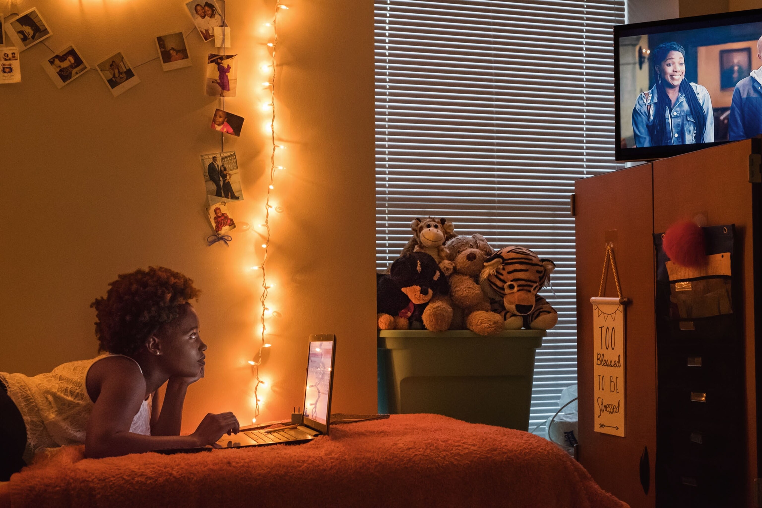 a young black woman in her dimly lit dorm room watching tv as she lay flat on her bed