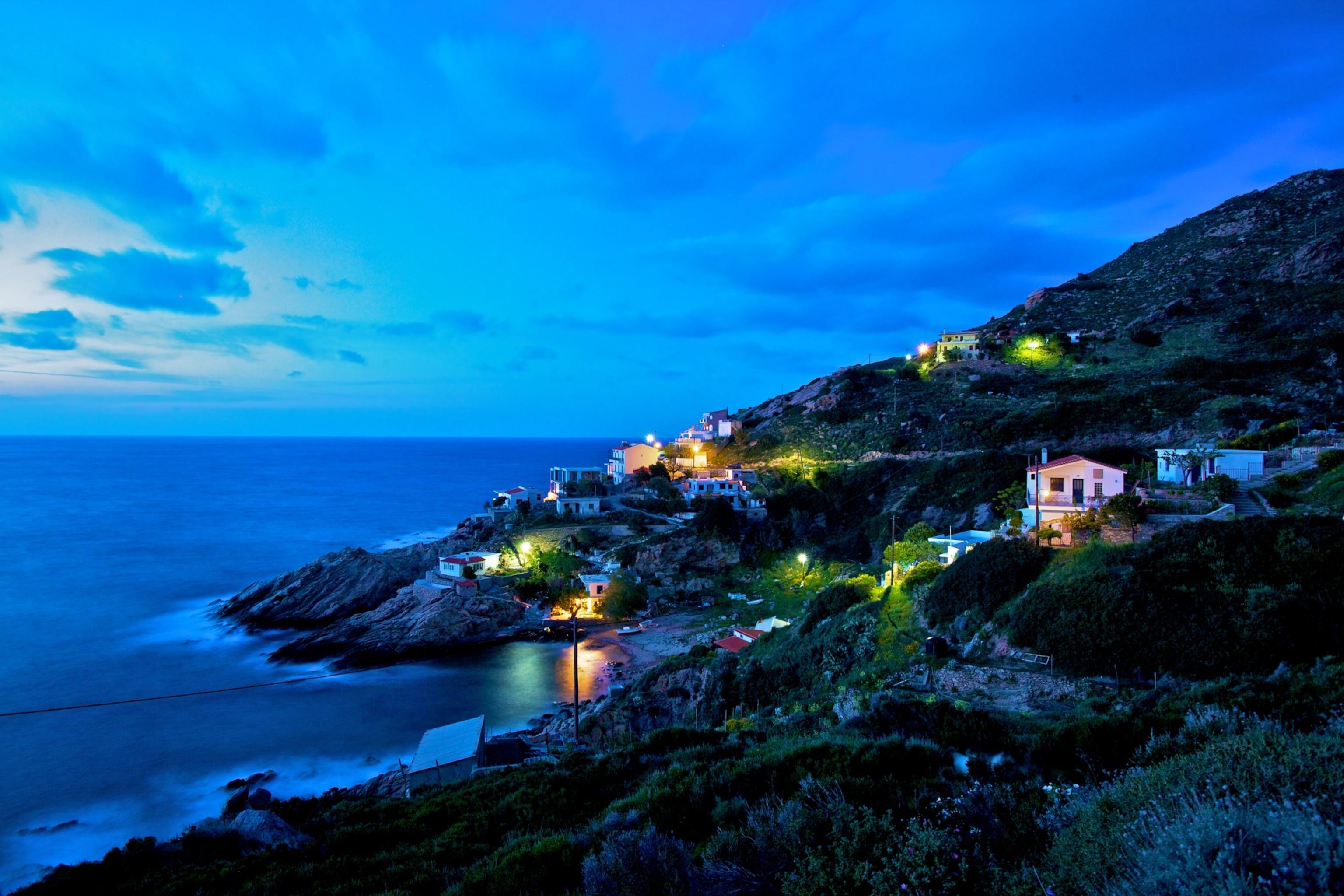 Several modern homes glow along the shore of Ikaria Island at dusk.