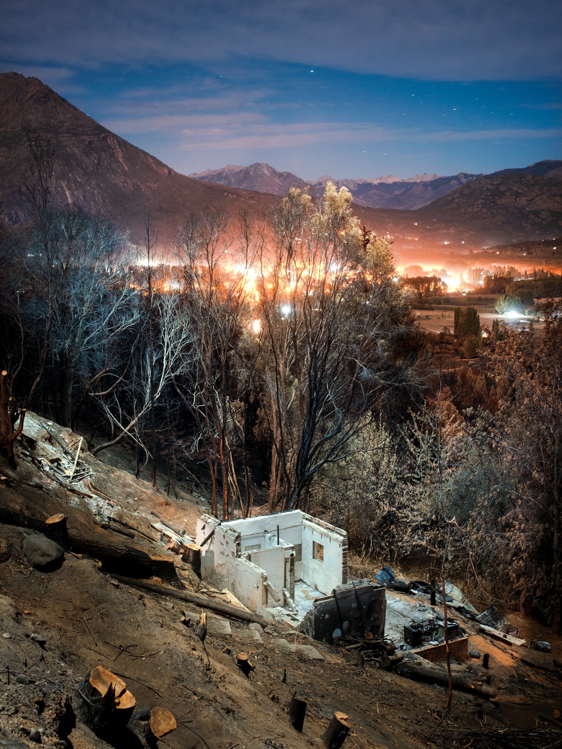 the charred remains of a home on a hill side with the lights of town glowing in the distance