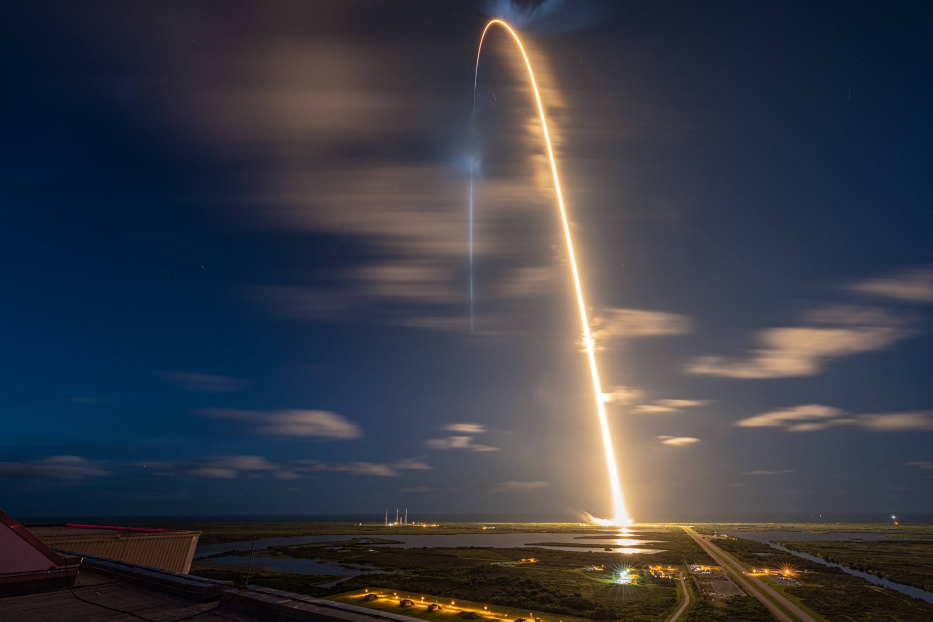 A long exposure of Inspiration4 launching, which appears as a golden arch of flaming light starting from the lower frame, extending upwards and away from the camera's perspective, out towards the ocean in the distance.