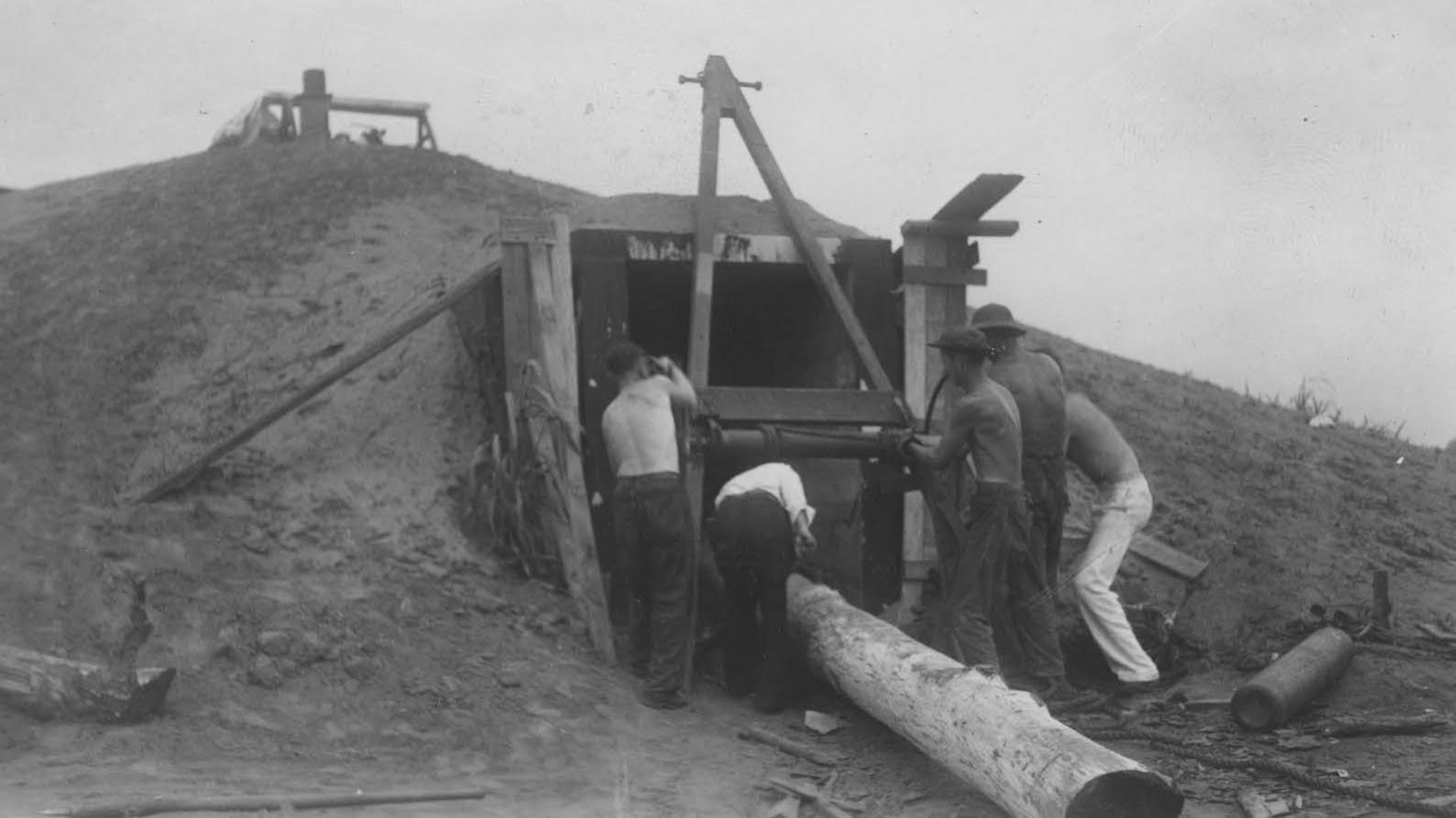 a black and white historical photograph of 5 males reconstructing the earth lodge. They are facing the lodge, maneuvering a large tree trunk, which is halfway inside the lodge, with a rope system,.