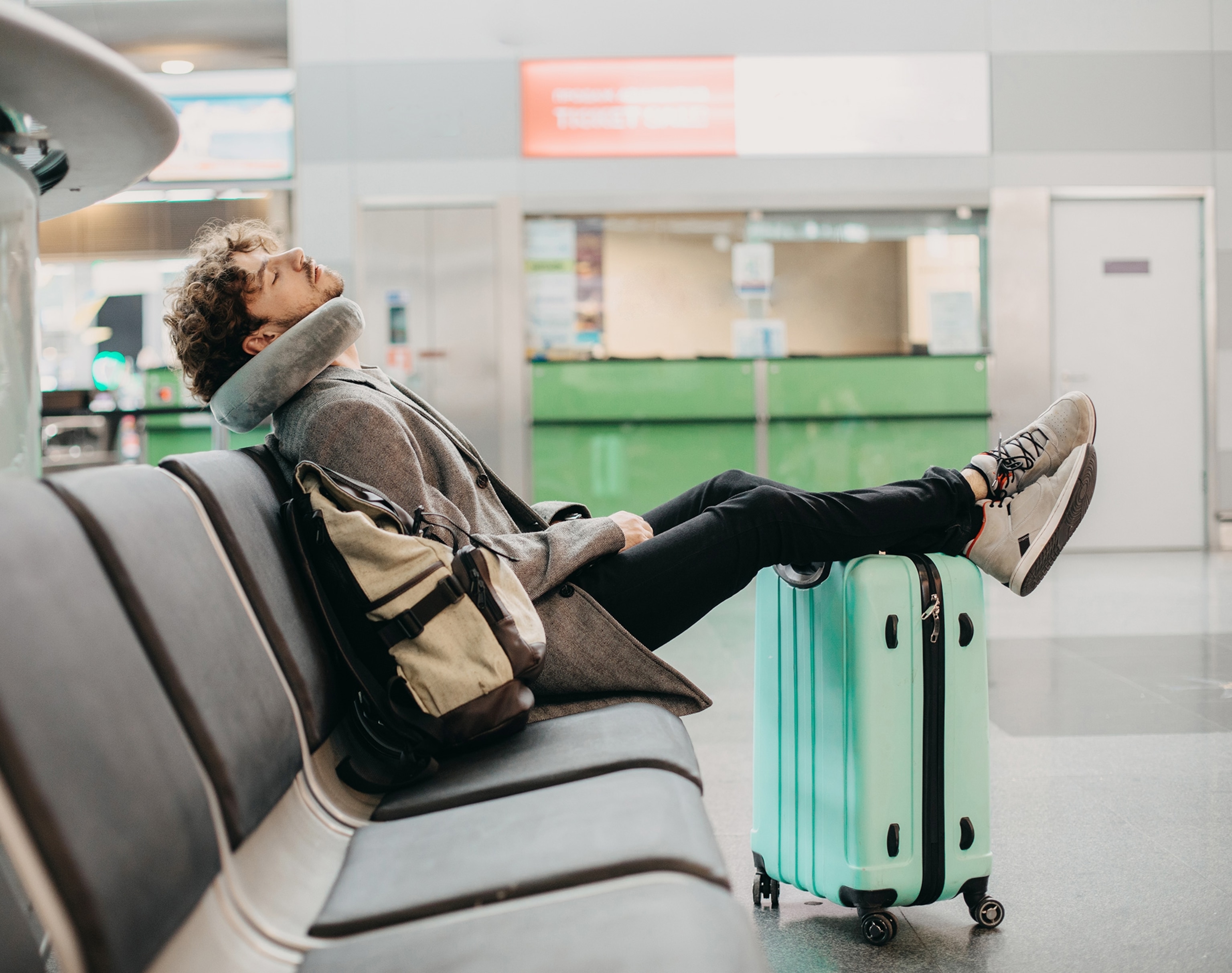 Young businessman sleeps alone in the waiting room of a station. He sits with his legs on suitcase and a pillow around his neck.