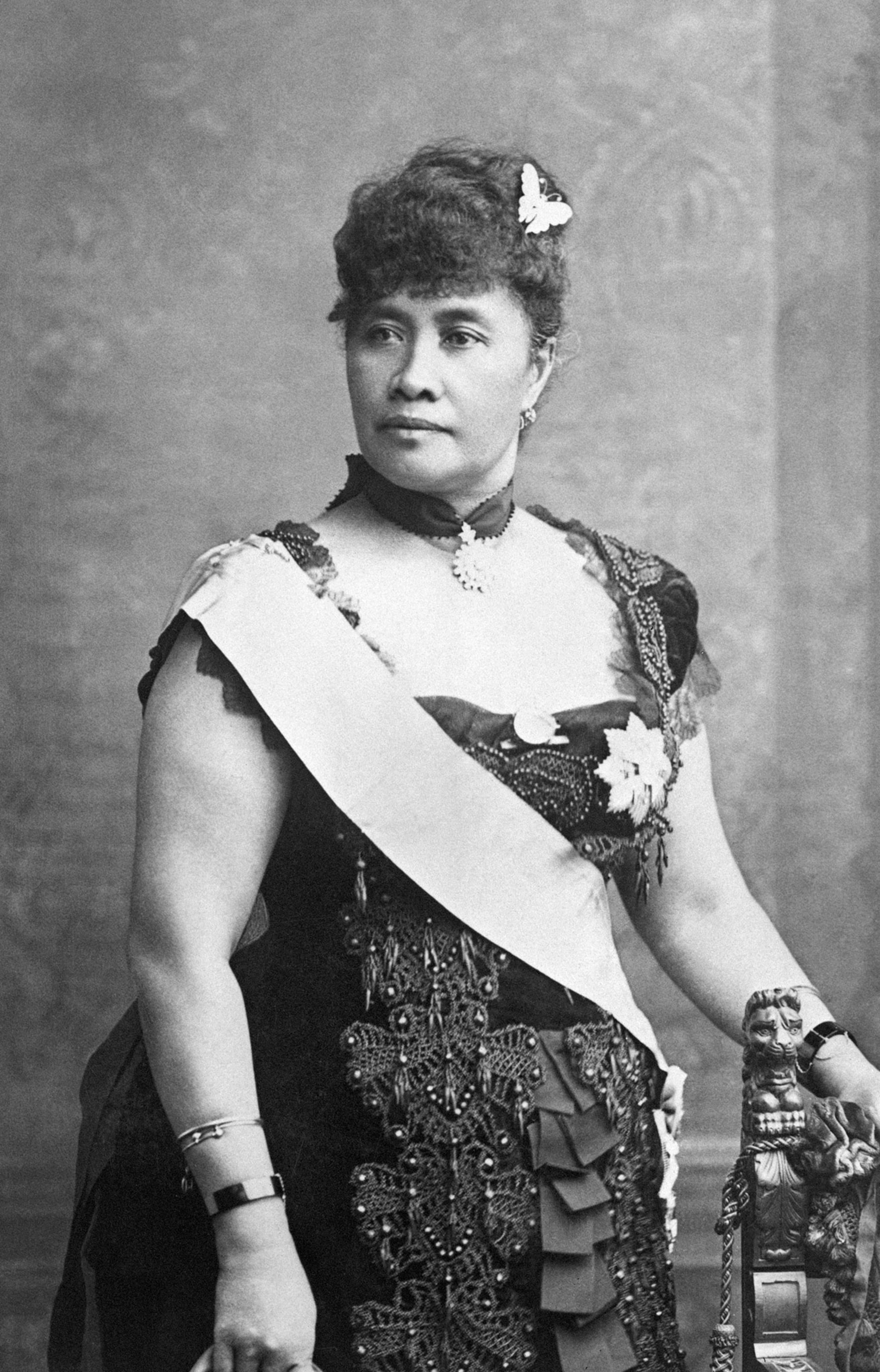 A black and white photo of a woman, dressed in an 1880s style dress with a sash drapped across her upper body. She is also wearing her hair up, with a butterfly clip, and has a thick chocker with an elaborate, silver-dollar-sized bead hanging from the center.
