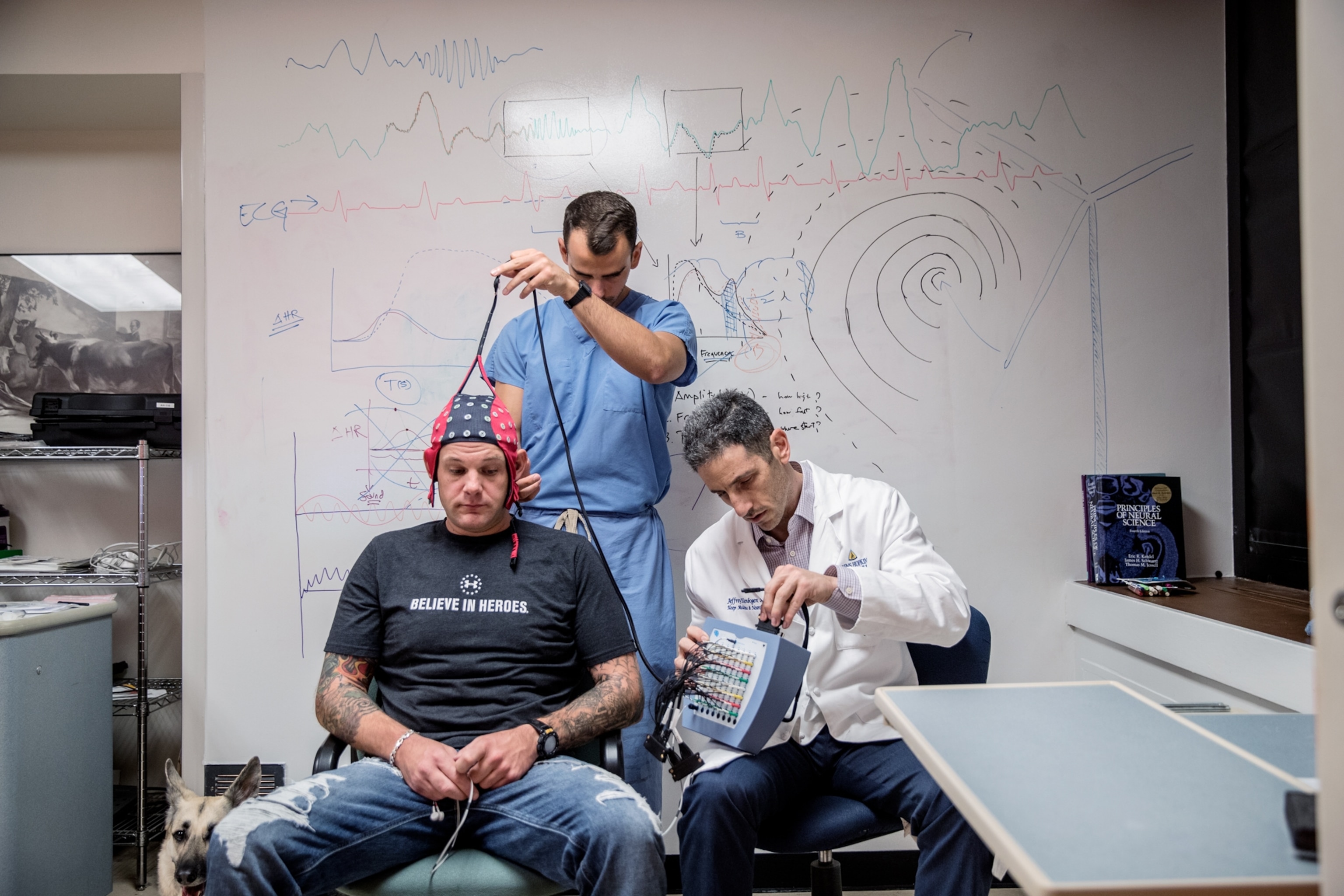 two men placing a EEG cap on a veteran as the veteran's dog sits beside him