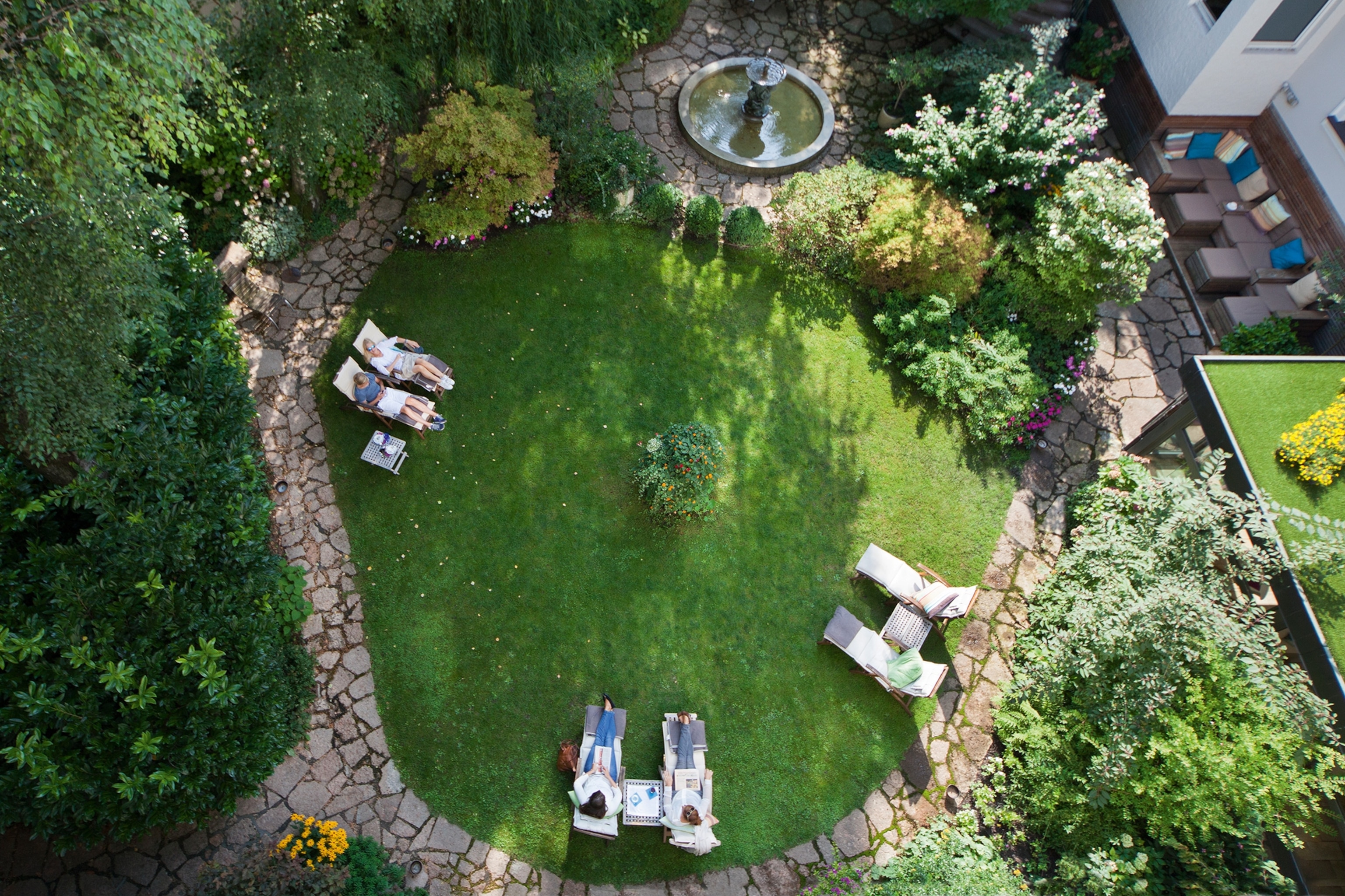 A lush garden from above, with a circling stone path, sunchairs and a small fountain.