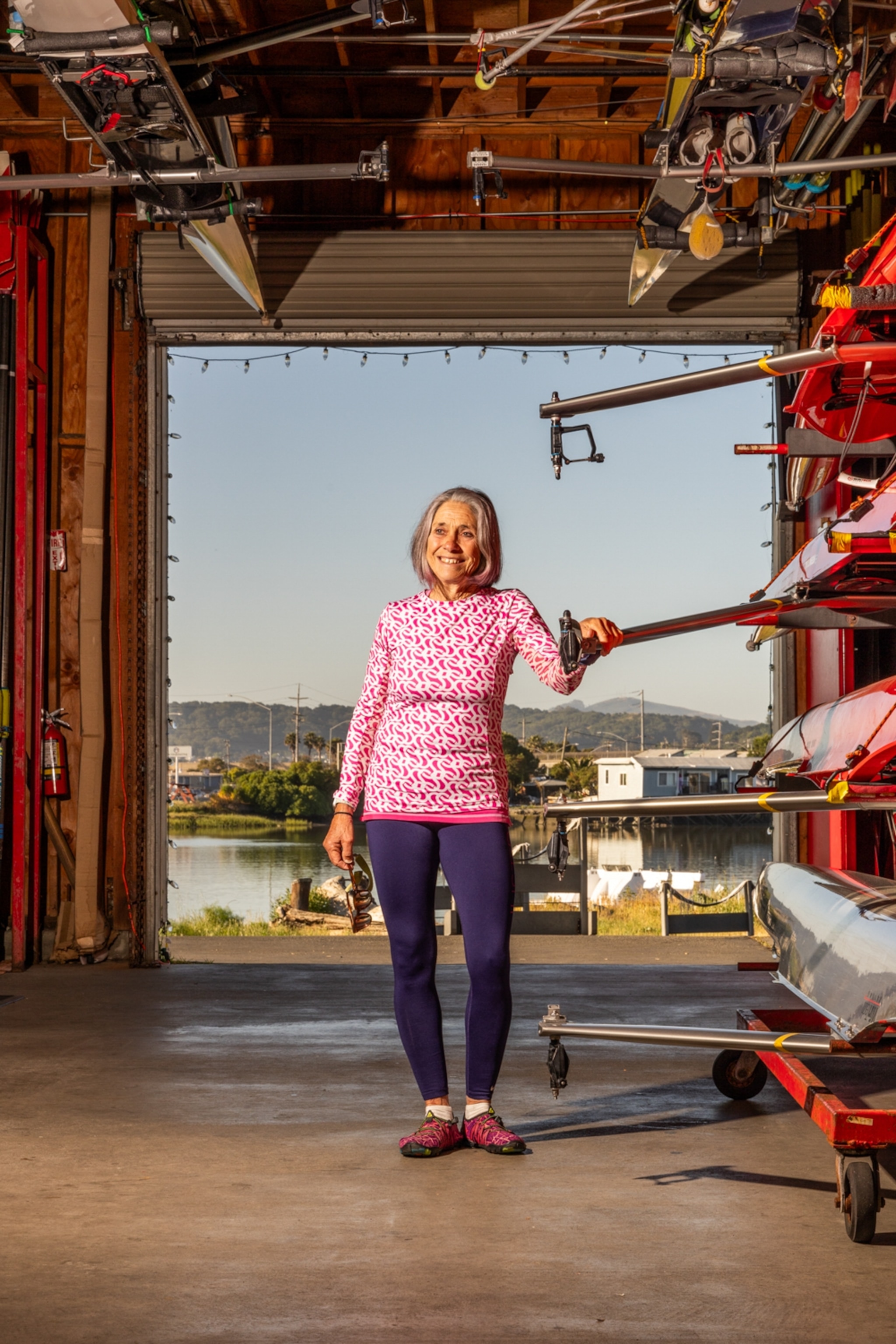 A woman stands in a boat house she is wearing a ping and white patterned top with purple leggings and a blue sky behind her.