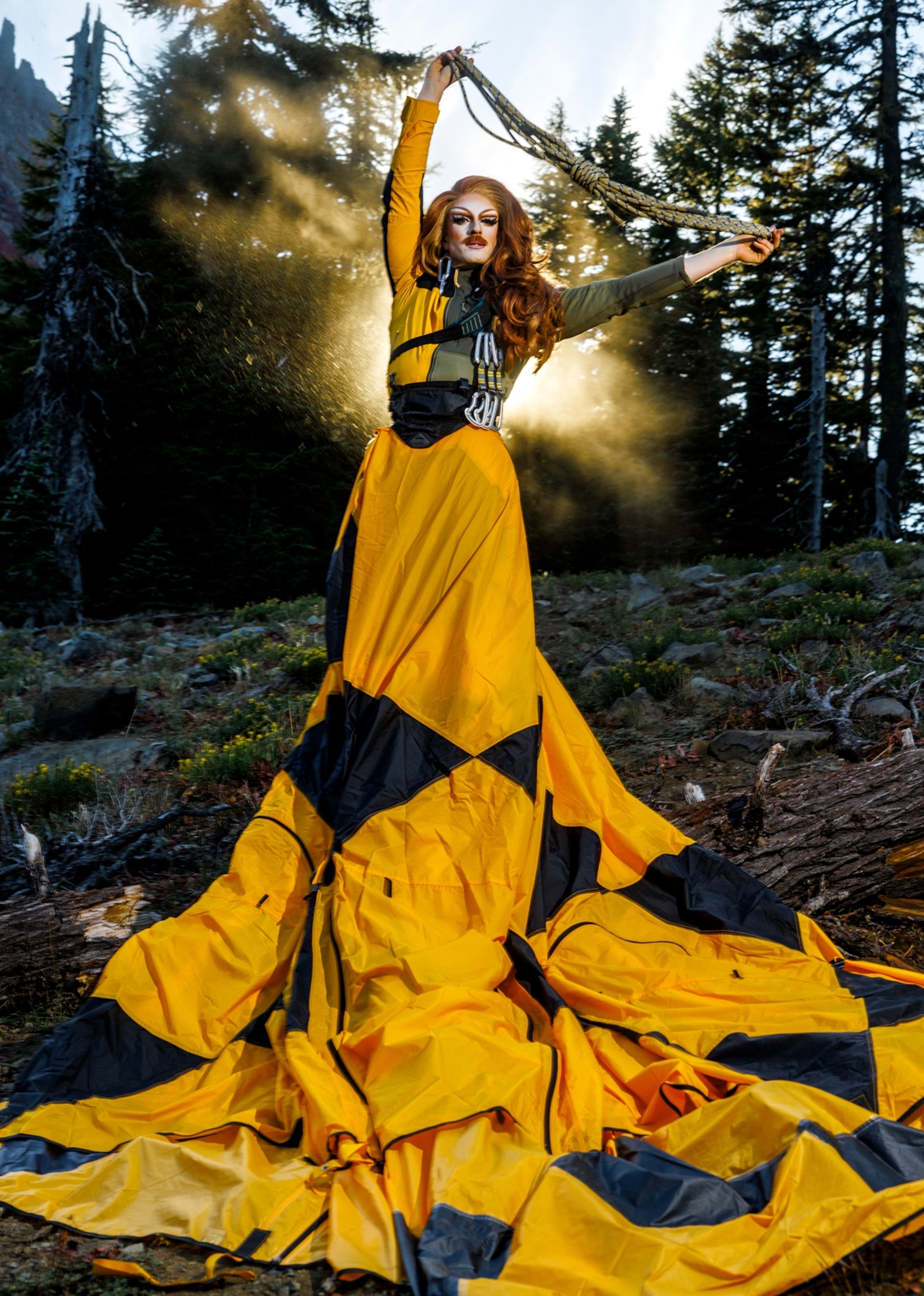 A person dressed in drag wearing a long yellow and black dress stands in the forest with the sun beaming from behind