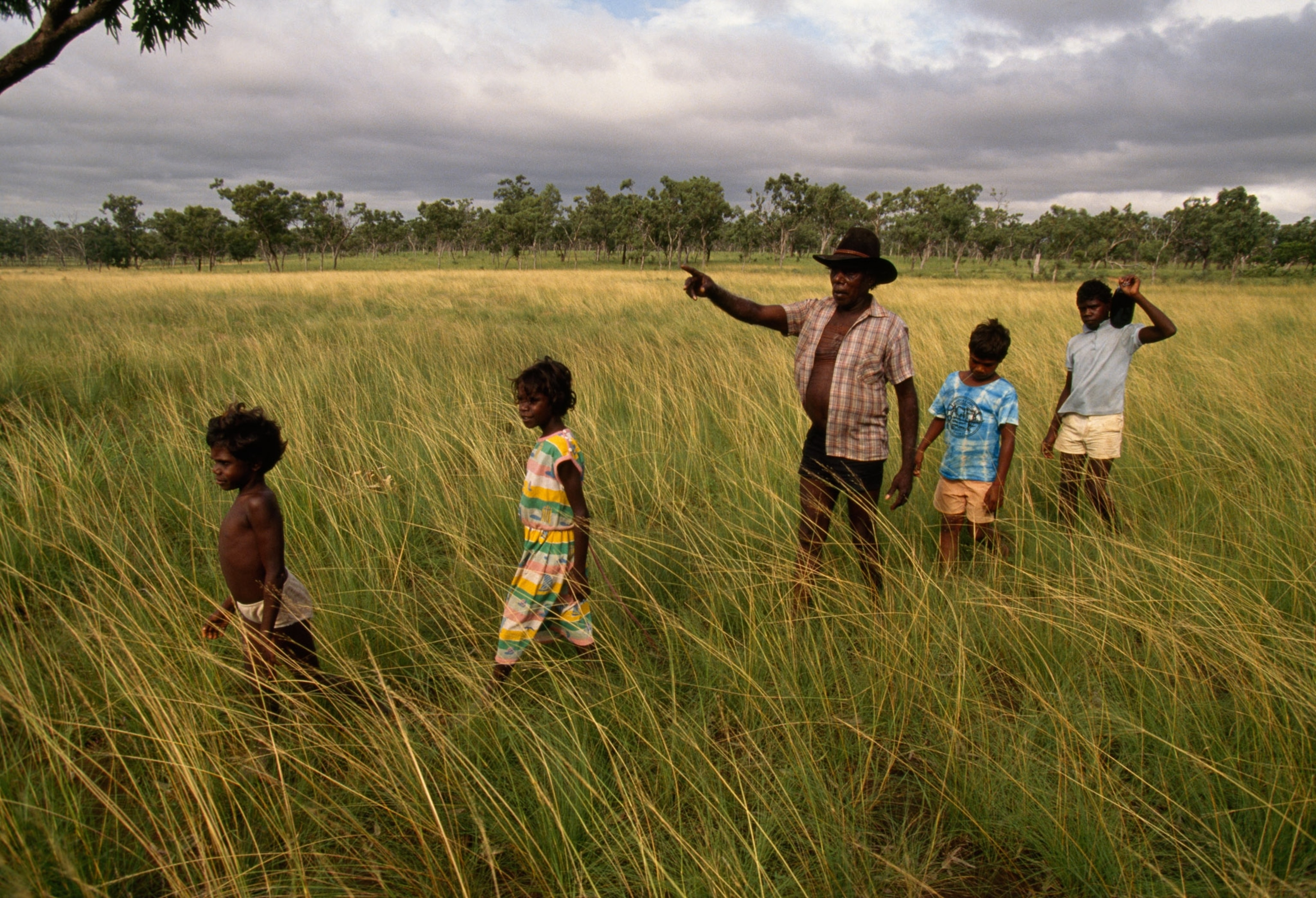 An older man with rich brown skin walks with children through a field point towards the sky.