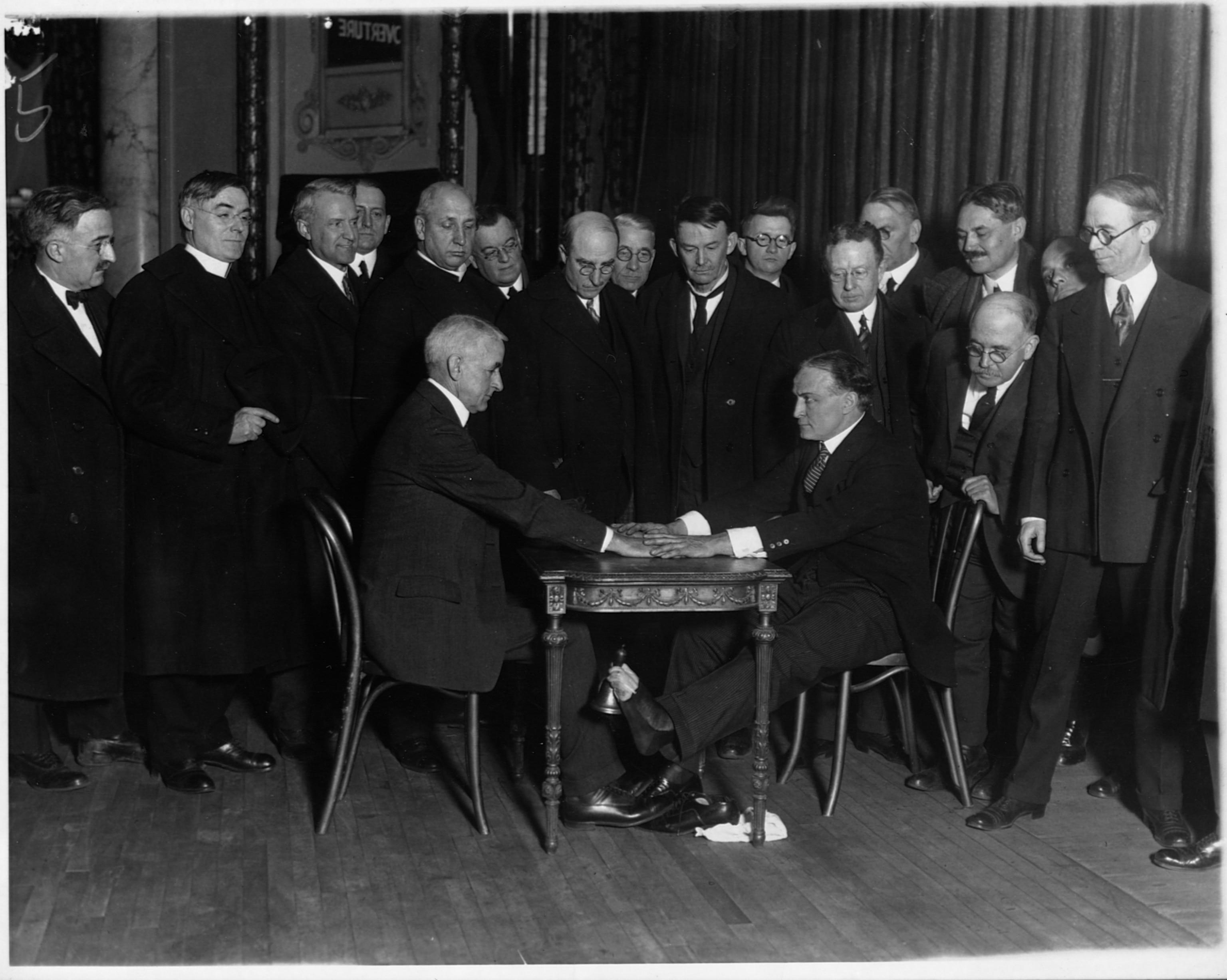 A group of men in dark suits standing around Harry Houdini, who is seated at a table with another man. Visible beneath the table, Houdini is using his feet to hold and ring a bell.