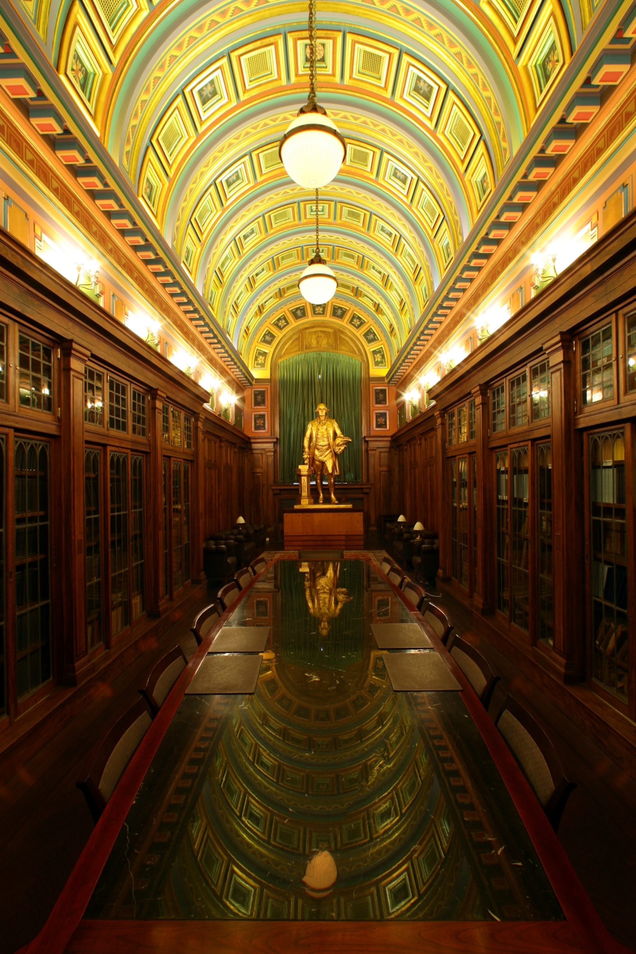 A long wooden table with a high polish panel reflects an ornate vaulted ceiling of a long narrow room.
