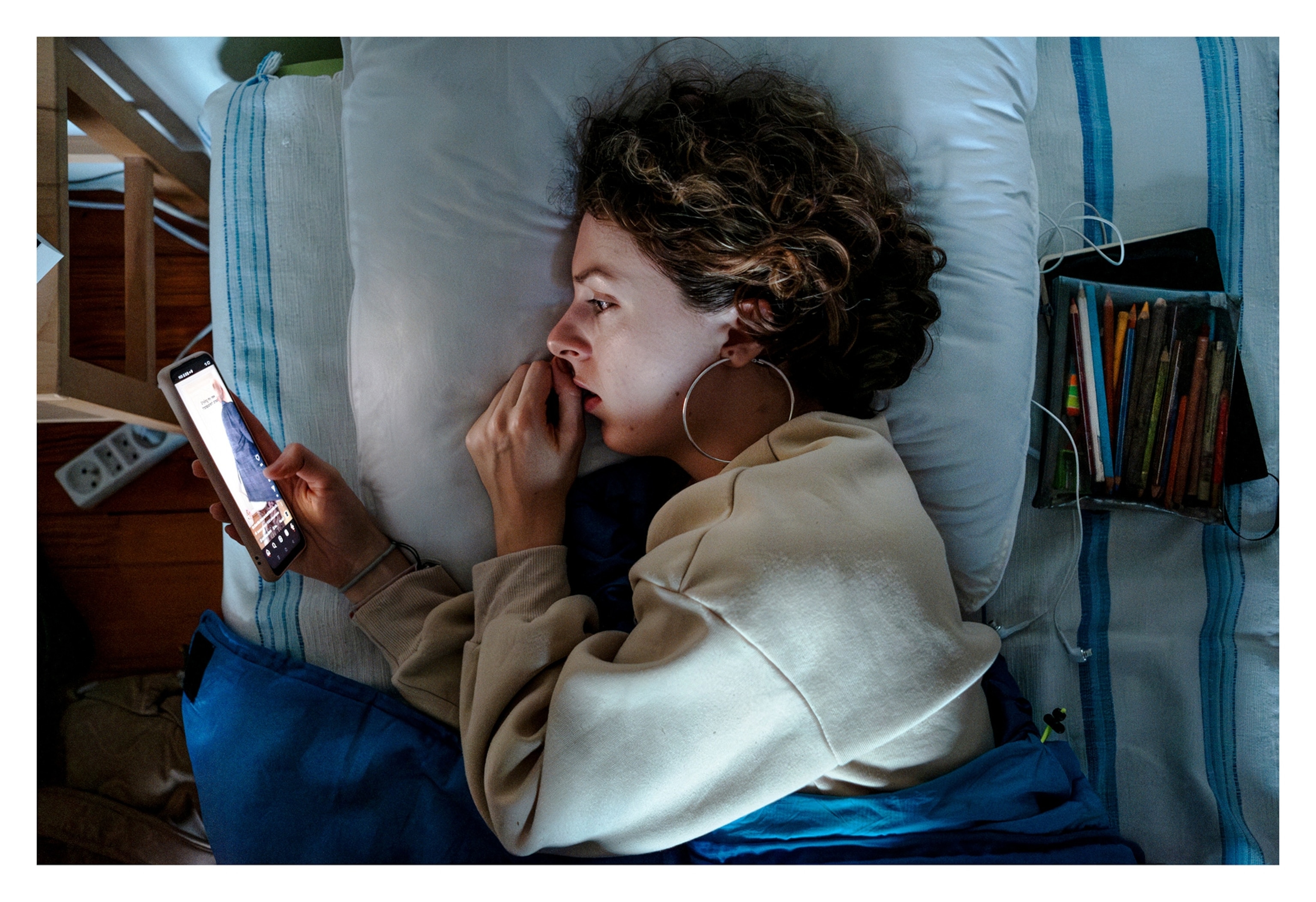 A young woman lays in bed scrolling on her phone.