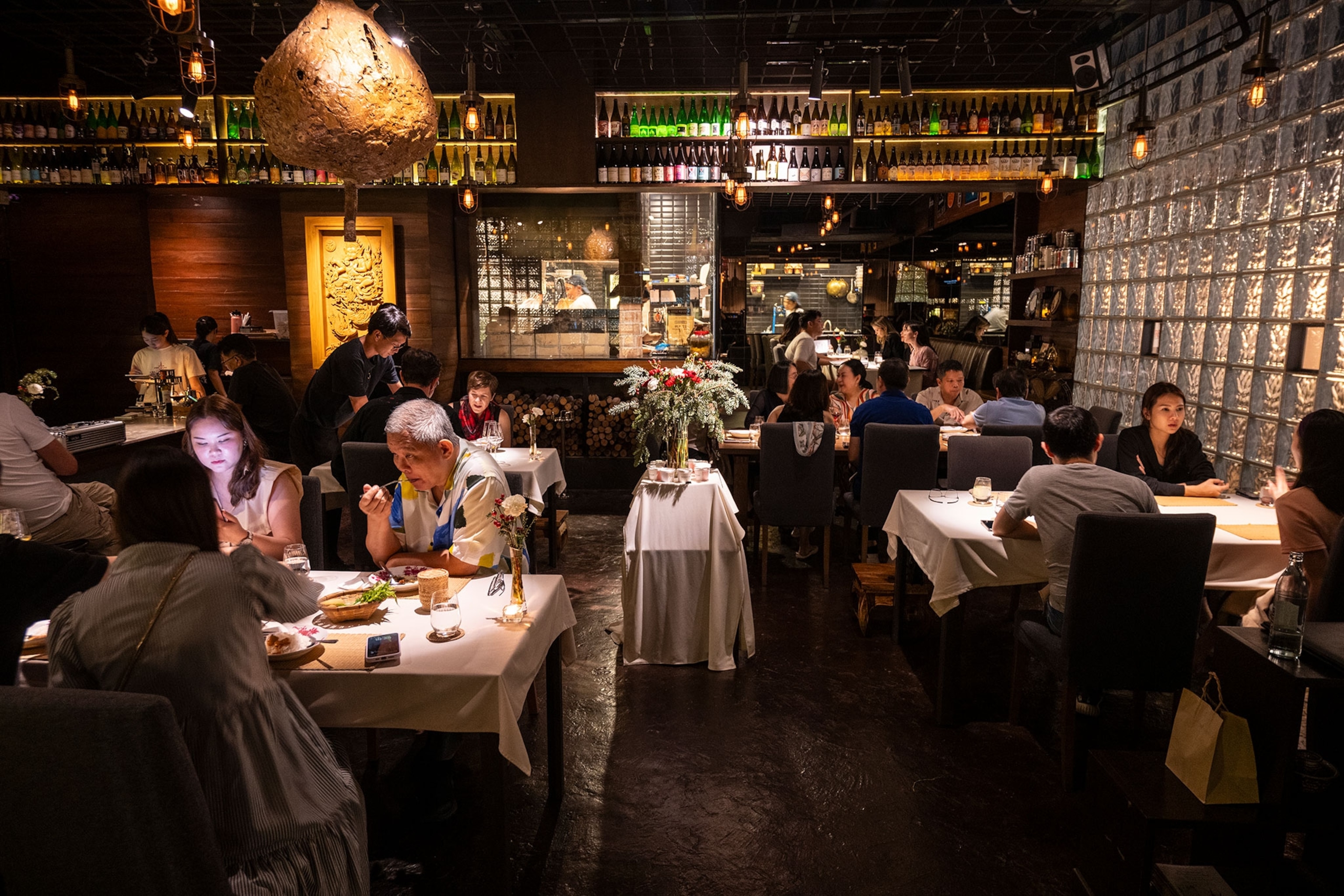 A darkly lit restaurant filled with people at tables lined with white clothes. The wall to the back is lined with bottles and a hive hangs from the ceiling as decoration.