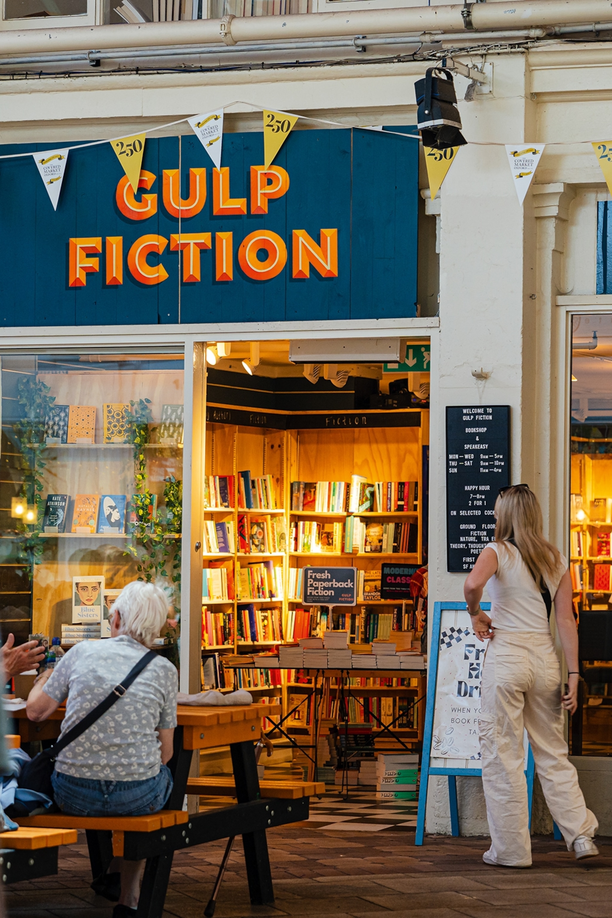 Girl standing at the entrance to a bookshop with overhead sign reading Gulp Fiction