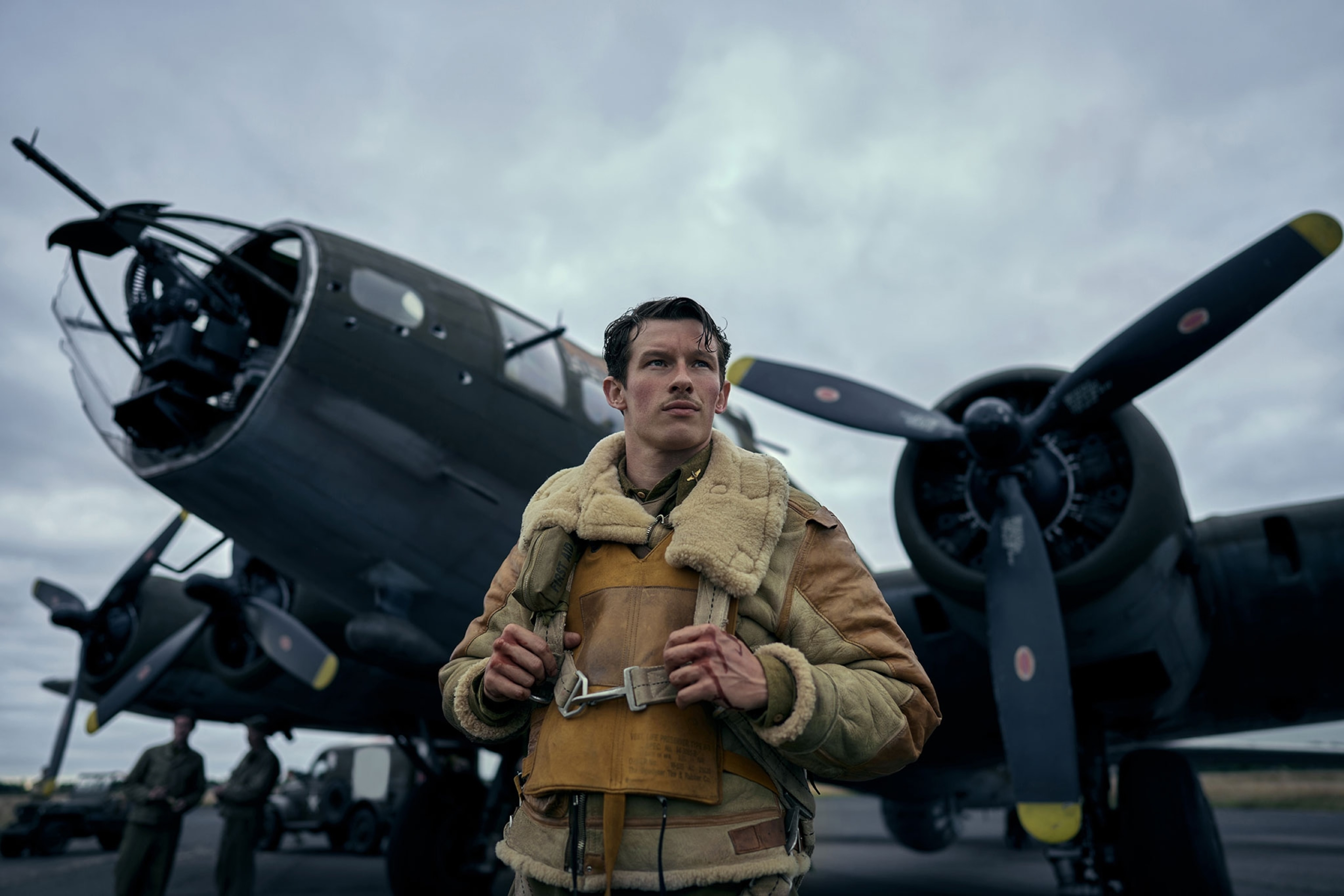 Callum Turner in "Masters of the Air,"as Major John Egan, stands in front of a dark, grayish-green, fighter jet with propeller engines. He is wearing a brown and tan leather bomber jacket that has a sheep's fur textured collar. Wrapped underneath his collar is a dirty yellow uninflated life jacket.
