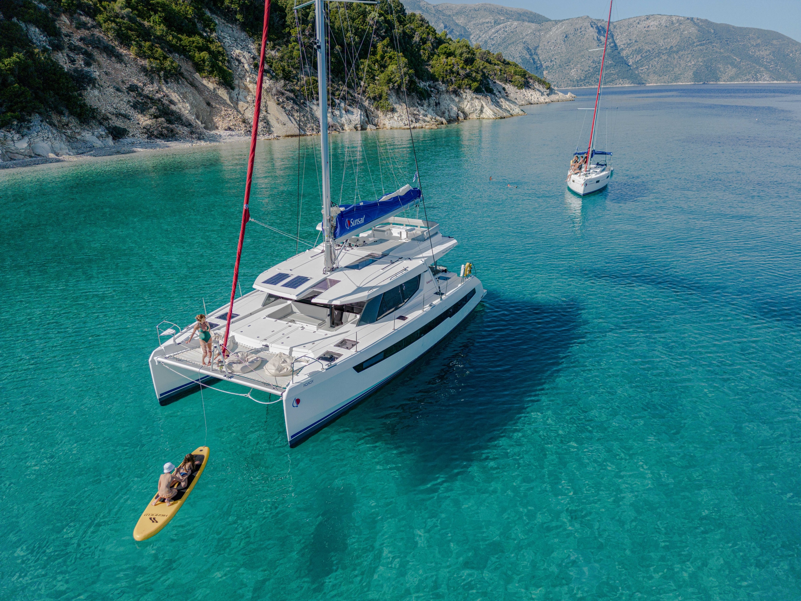 A catamaran and a sailboat are anchored in clear blue seas, with a sandy shoreline in the background. A man sits on a paddleboard in front of the catamaran.