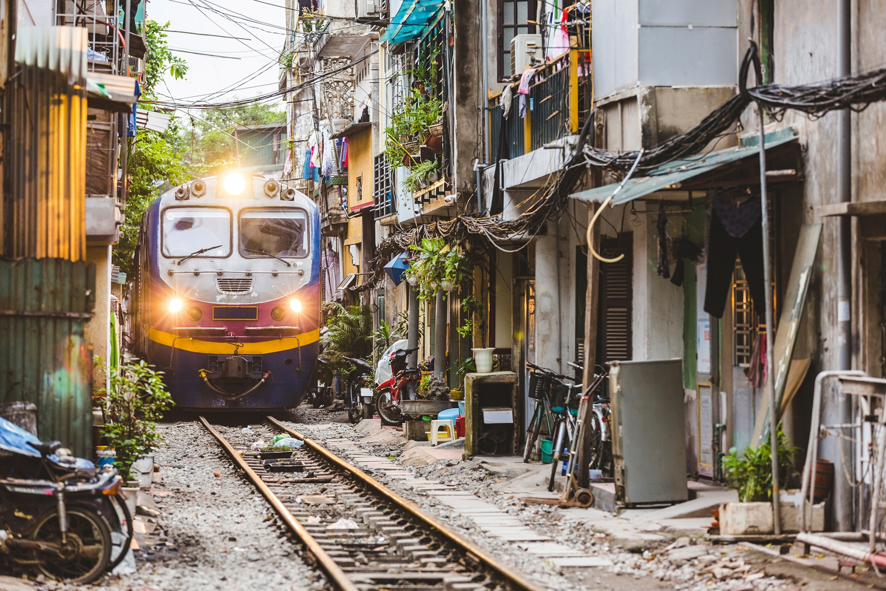 A train squeezing through a tight residential street with tropical plants and bikes along the road.