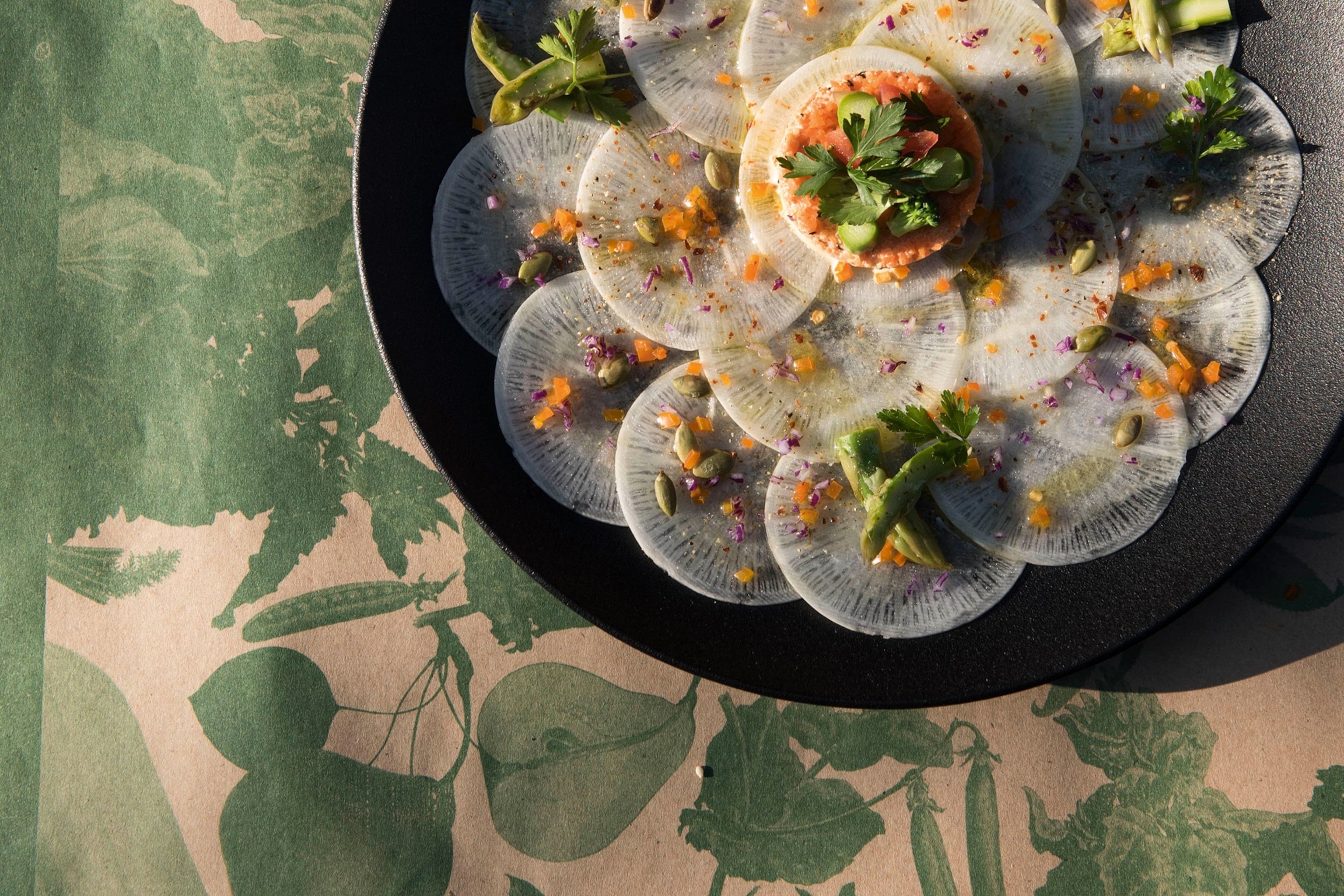 A black plate is sitting on a floral table with a dish of thinly sliced radish which is garnished with colourful herbs.