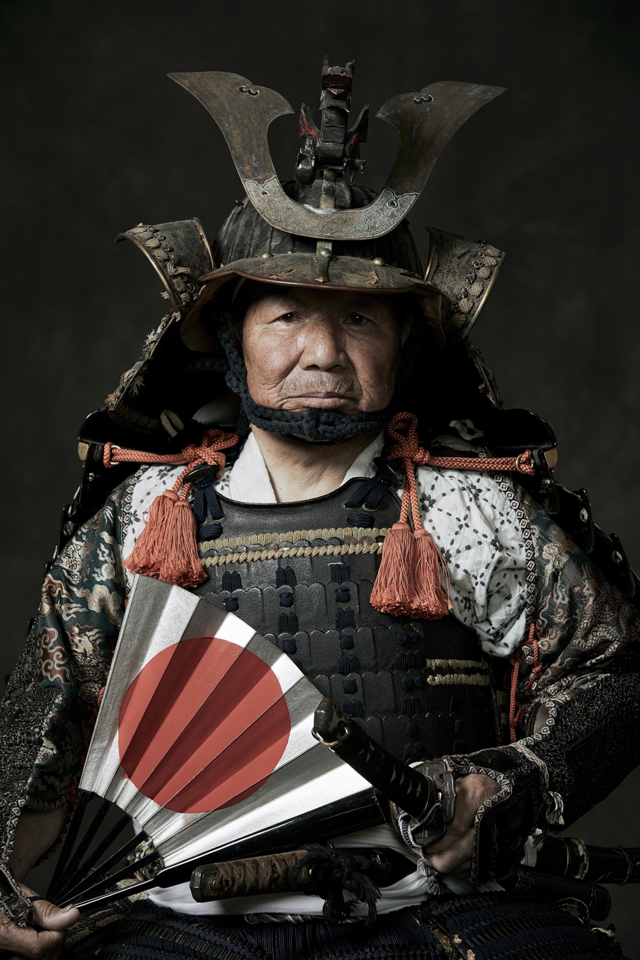 Picture of old man in large and complex helmet holding white paper fan with red circle on it.