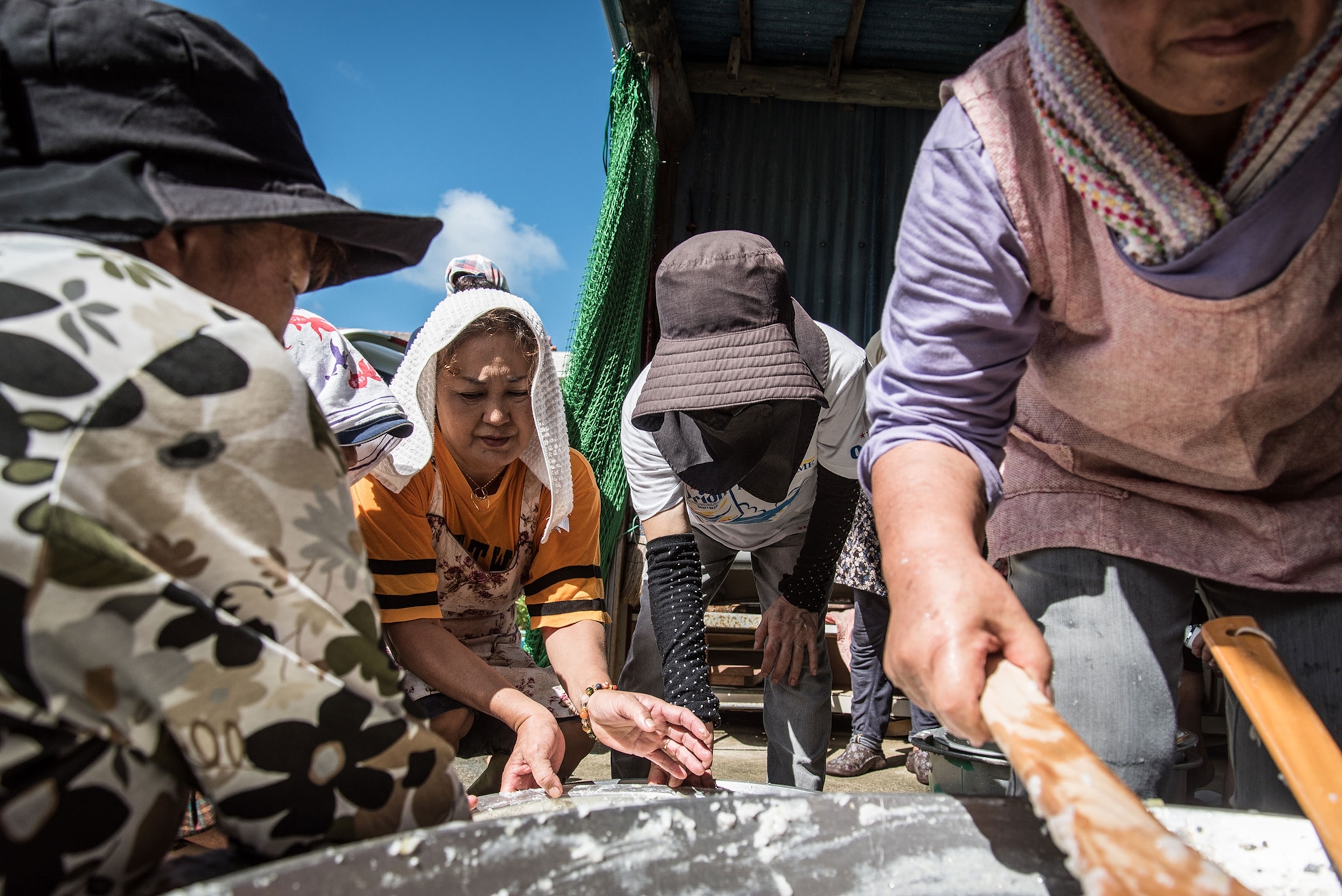 Okinawan women gather around large bowls preparing sake on a hot day. To protect from the sun, they wear hats and towels on their heads.