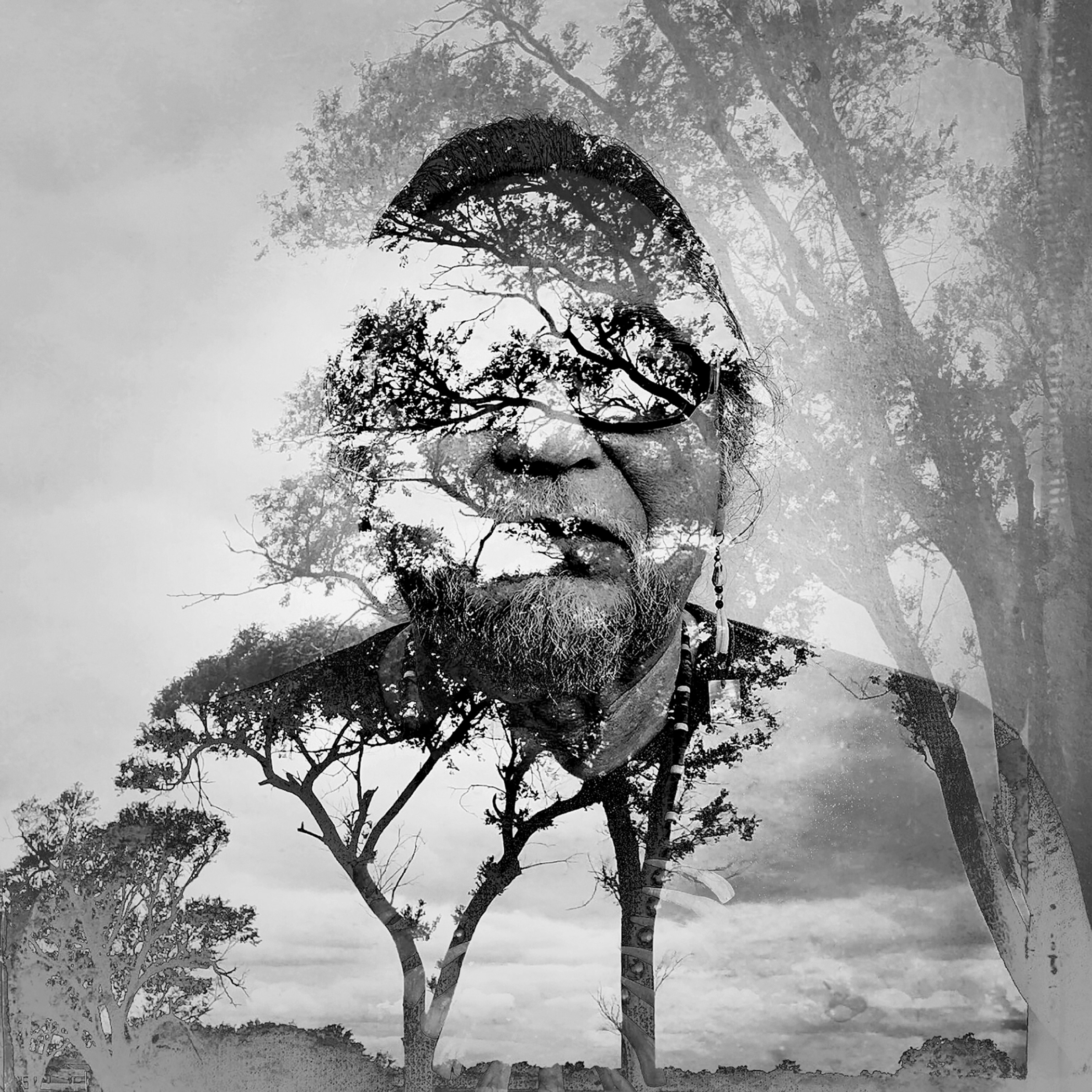 Picture of b&w portrait on a man with beard and eyeglasses and trees on background.