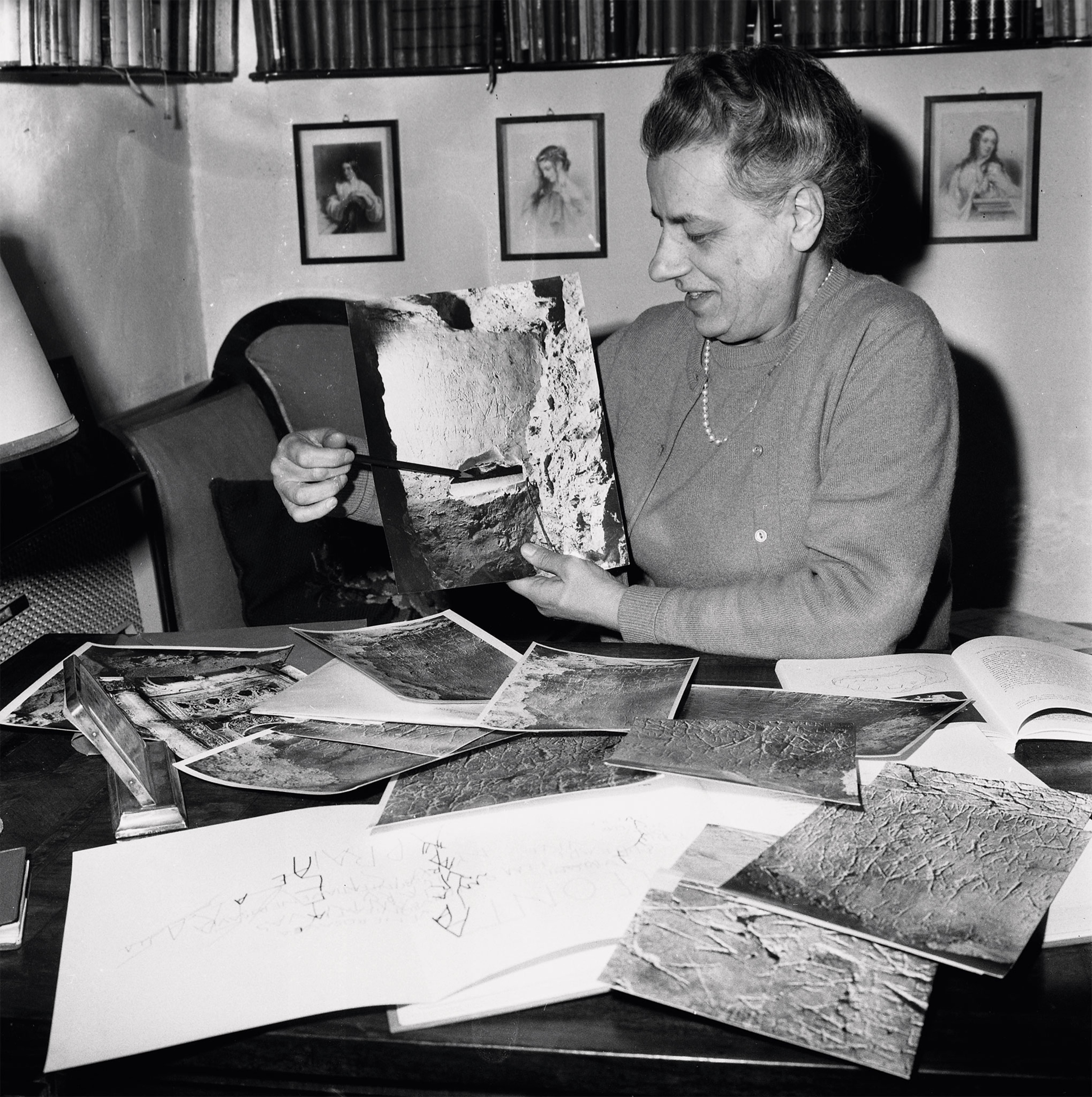 A woman points out the cavity in the tomb in a photograph, while sitting at a table covered in photographs
