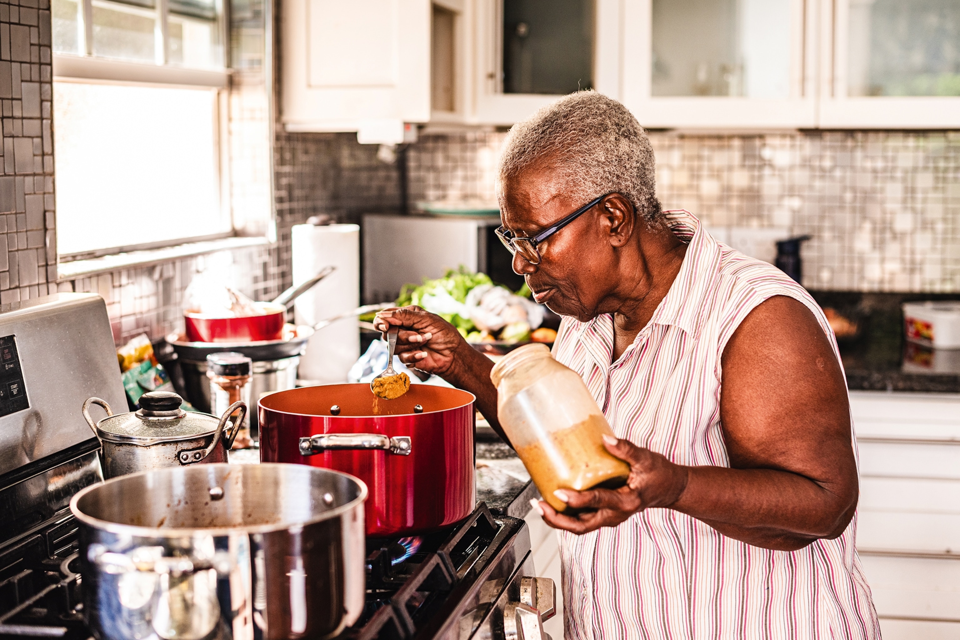 An older woman seasoning her stew in a pot on the hob with a spice powder.