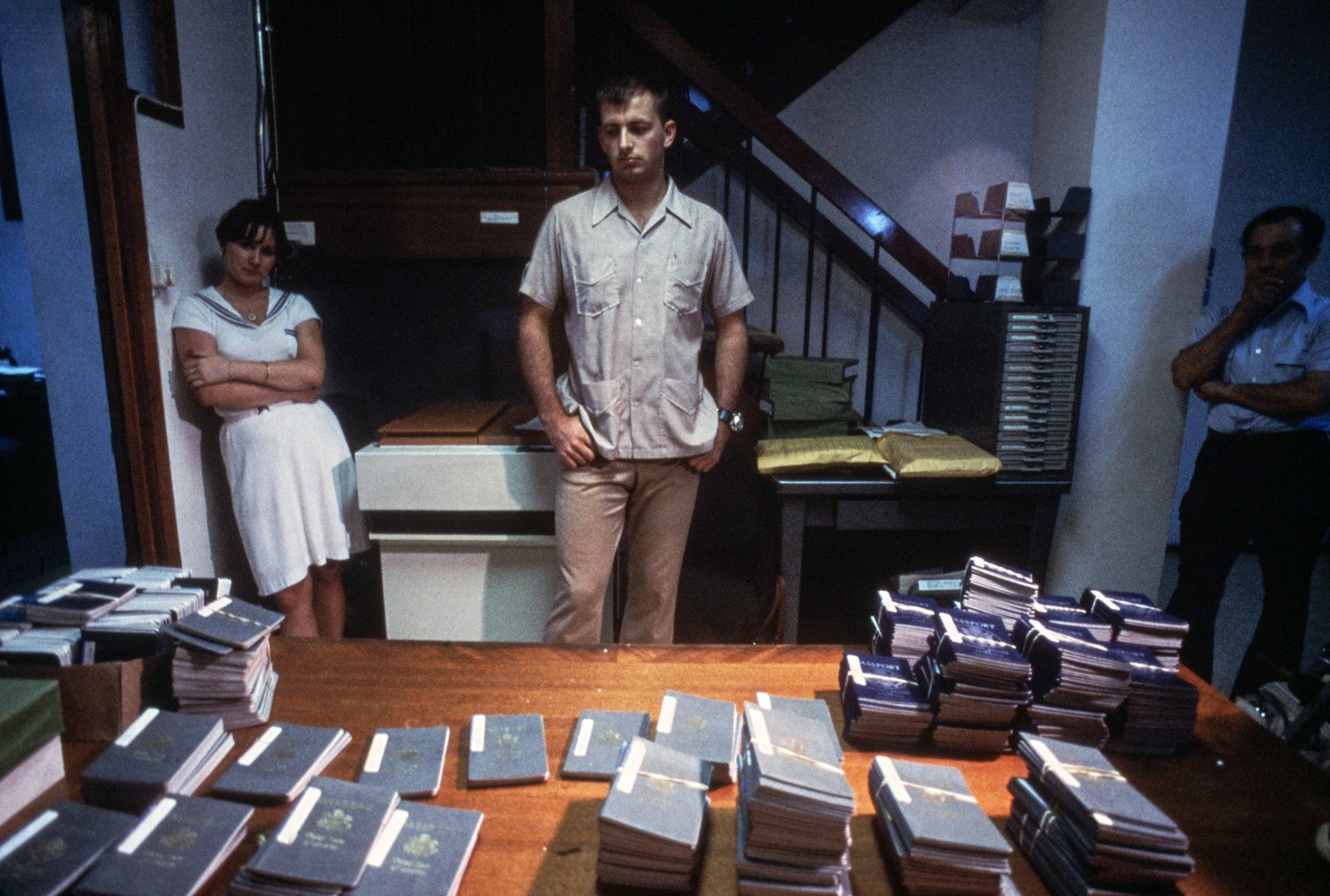hundreds of passports on a table with three people in the room looking at them.