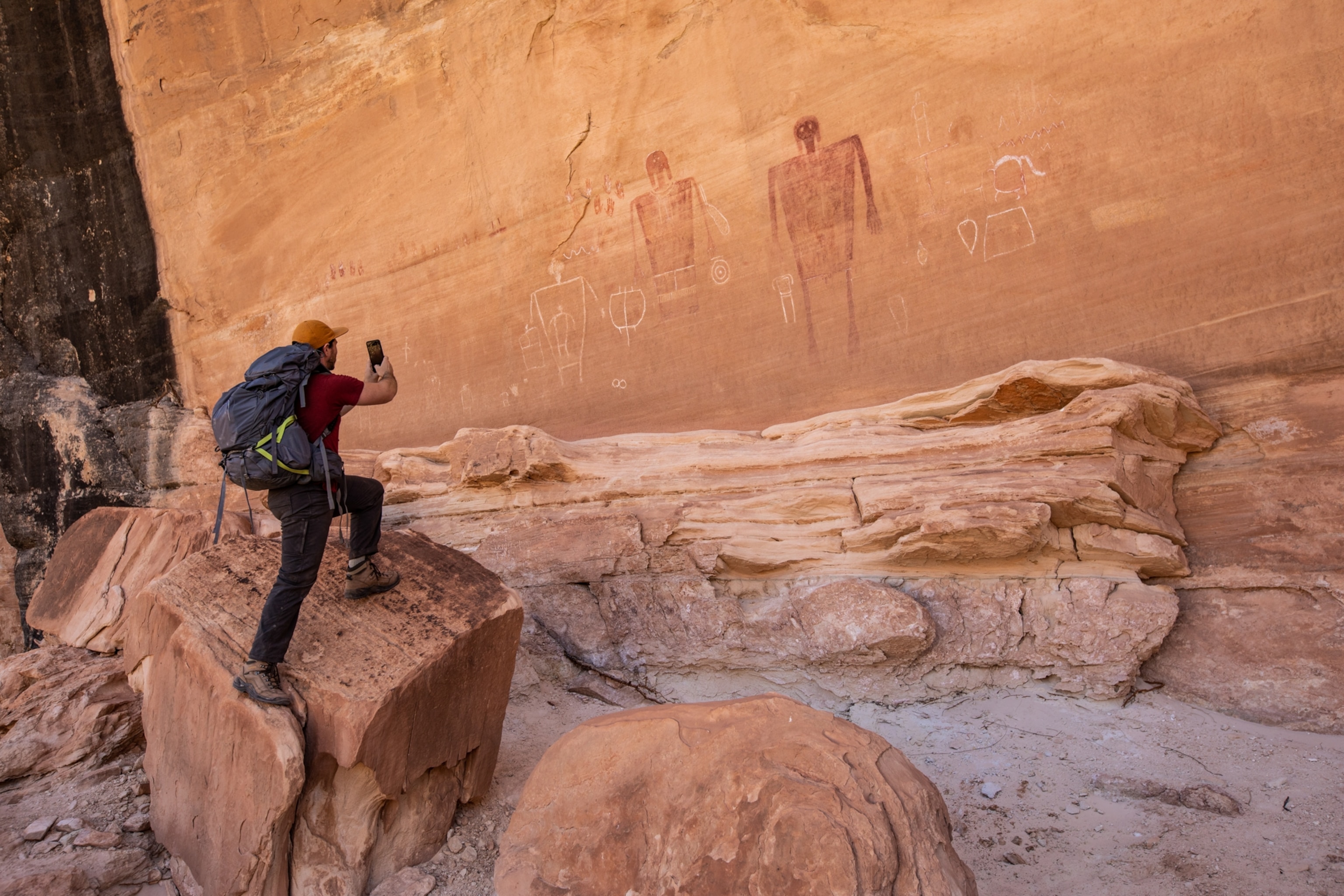 A man using his phone to photograph ancient cave art on a cliff wall depicting a human of similar height to the photographer.