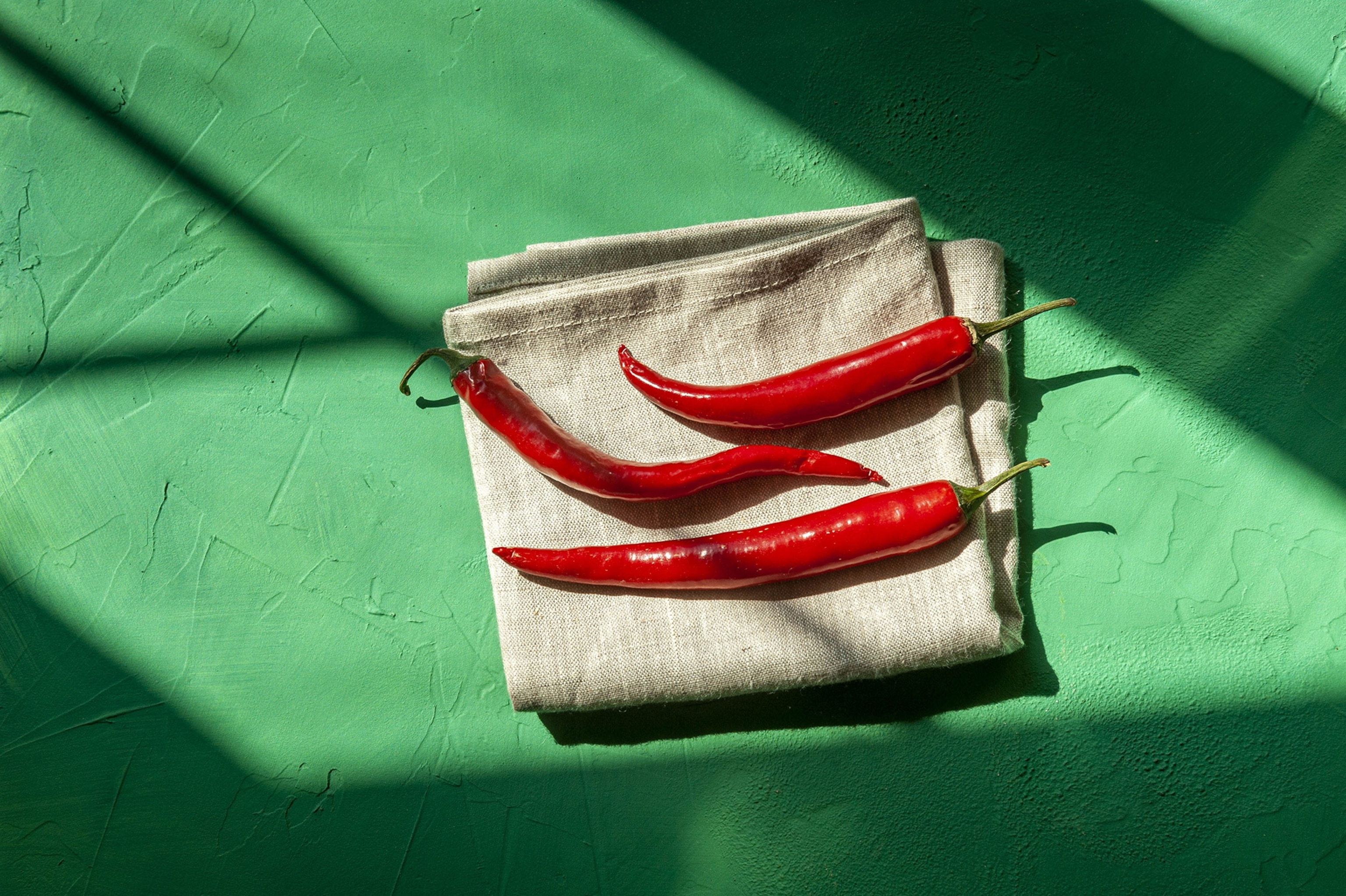 A close-up aerial view of three red chili peppers placed on folded linen with a vivid green textured surface beneath.