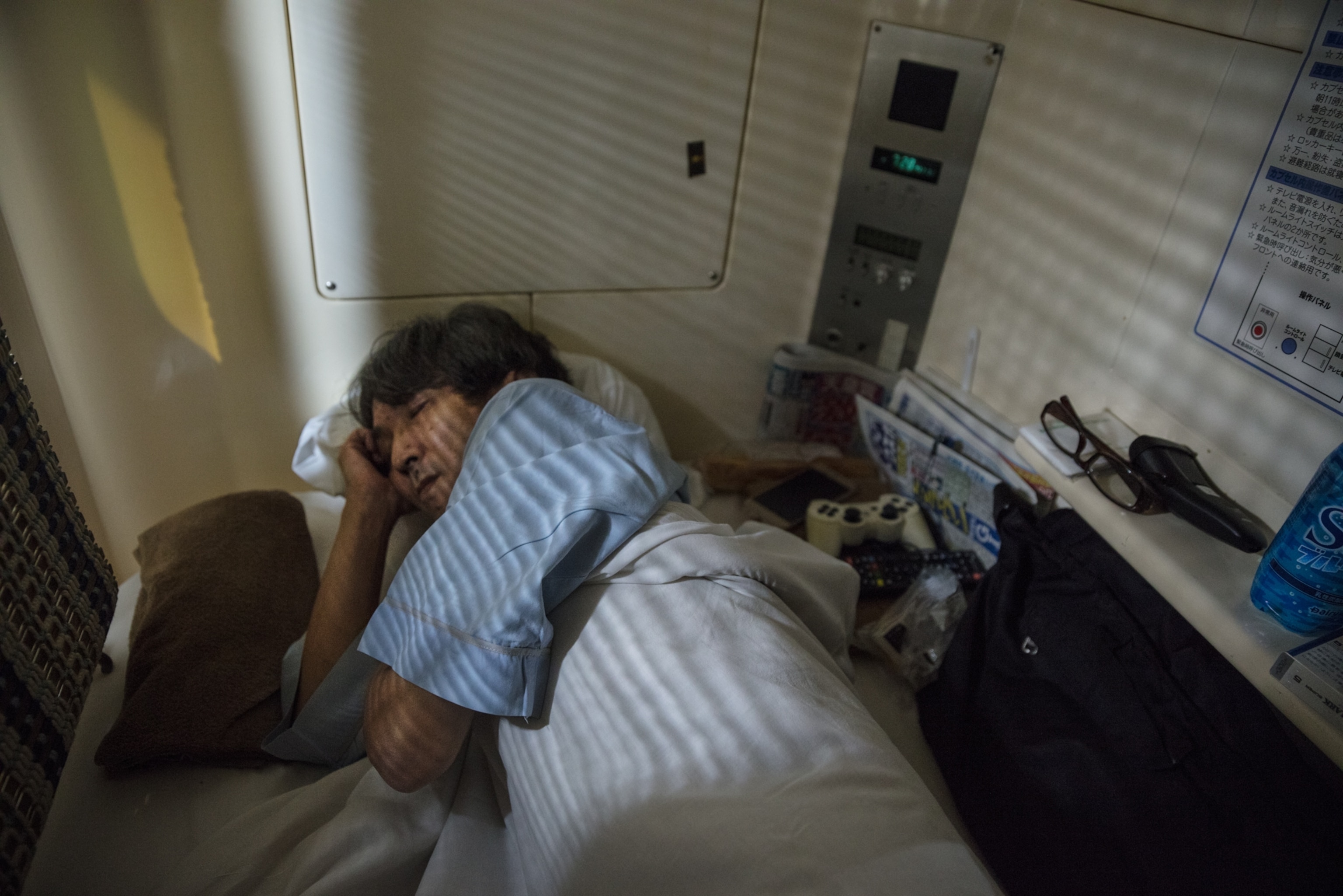 a man resting in his capsule hotel bed as the shadows of blinds rest on him