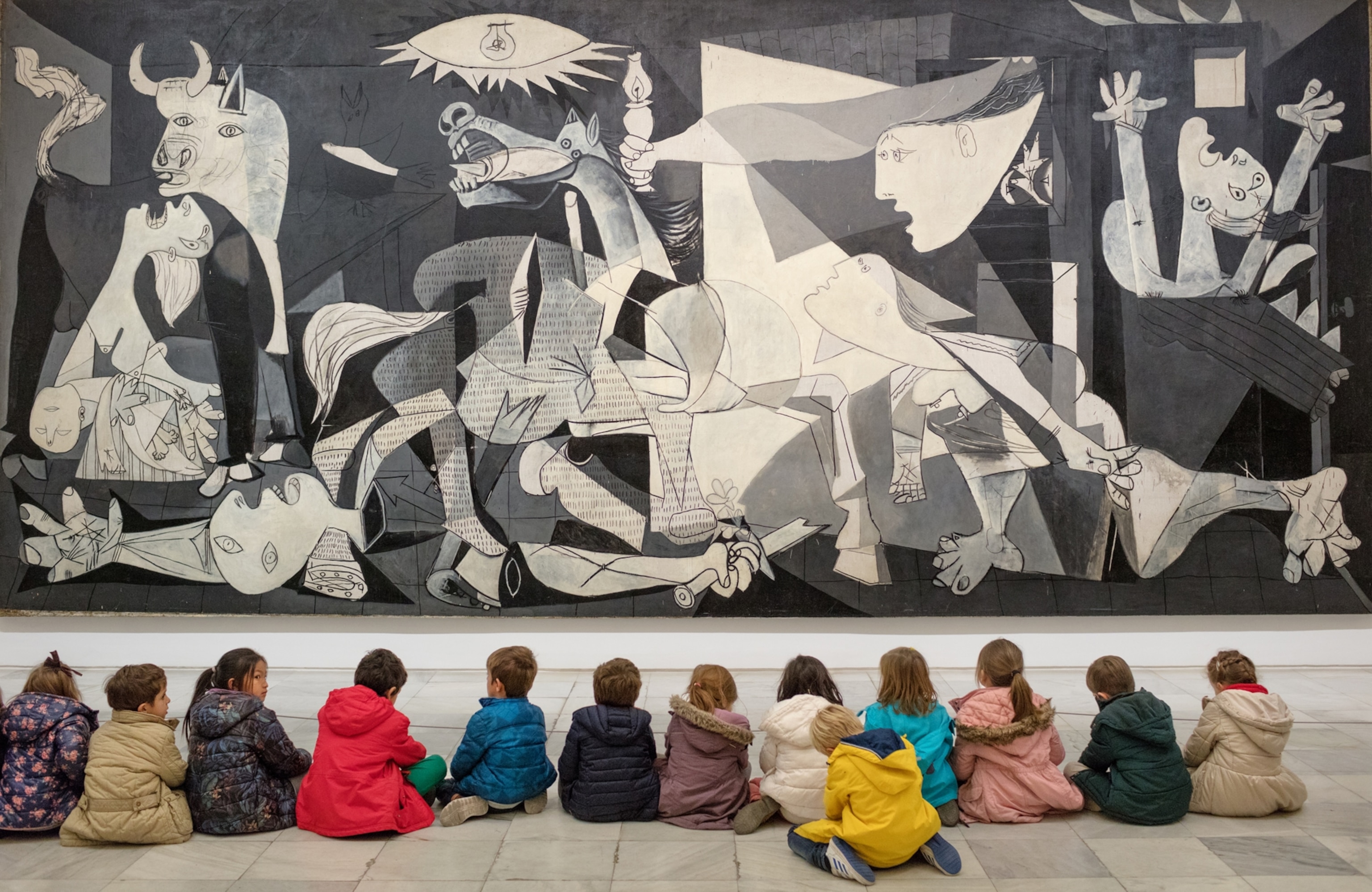 children sitting down in a horizontal line looking up at Picasso's Guernica