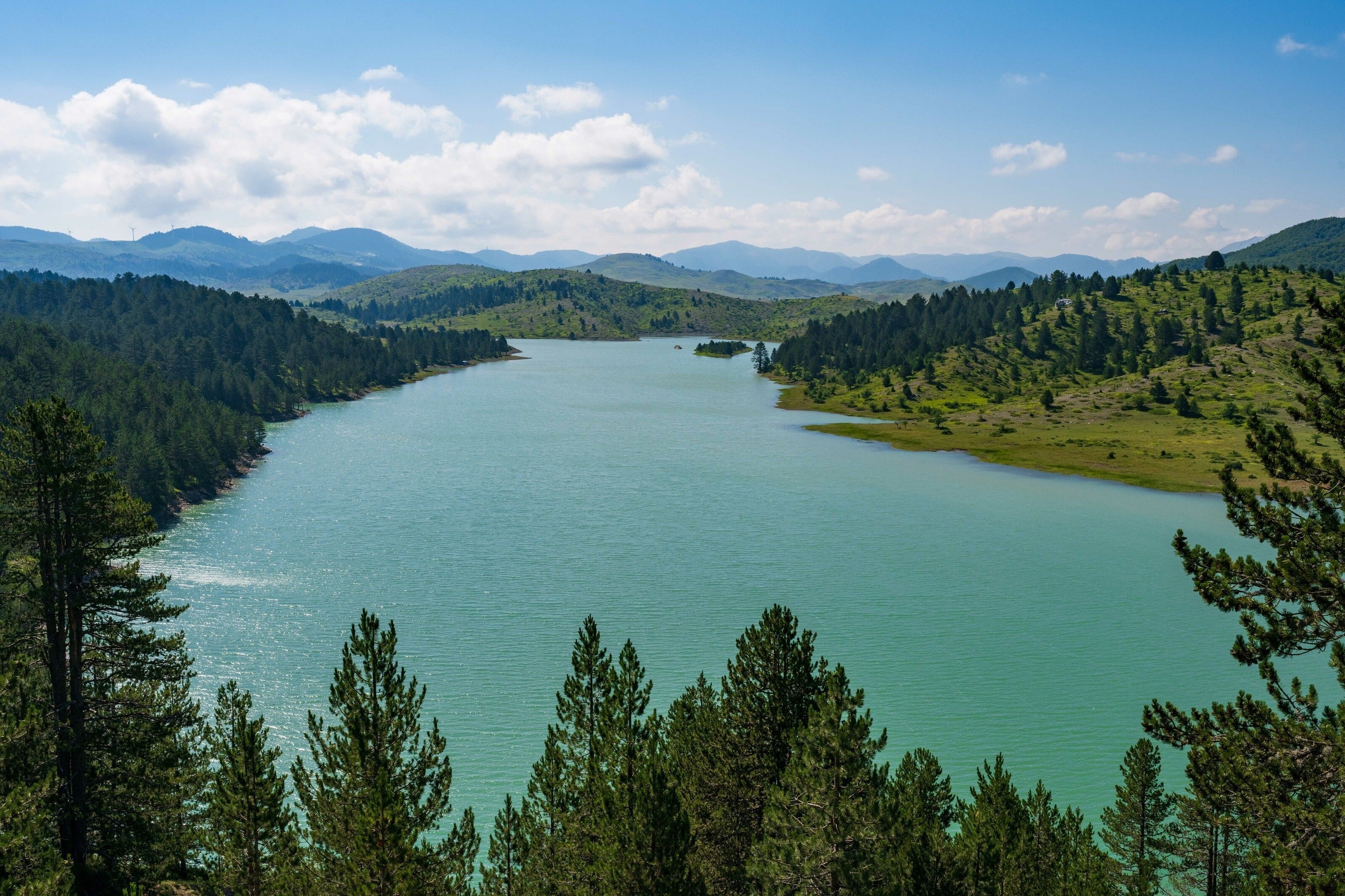 Lake Aoos, sat between pine-covered banks. Its waters are a cool turquoise, rippled by a mild breeze.