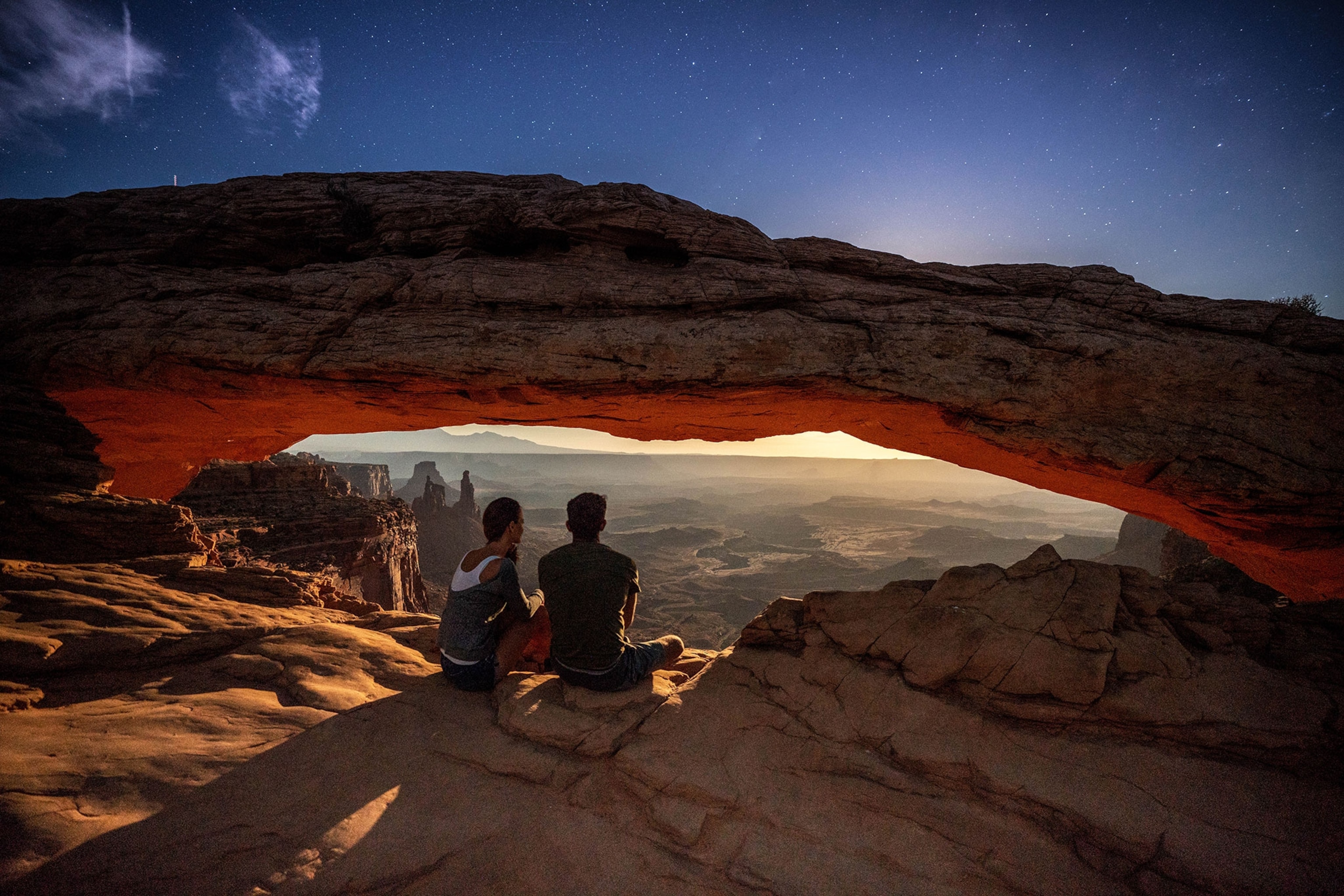 A couple look out on the rising moon under Mesa Arch at night.