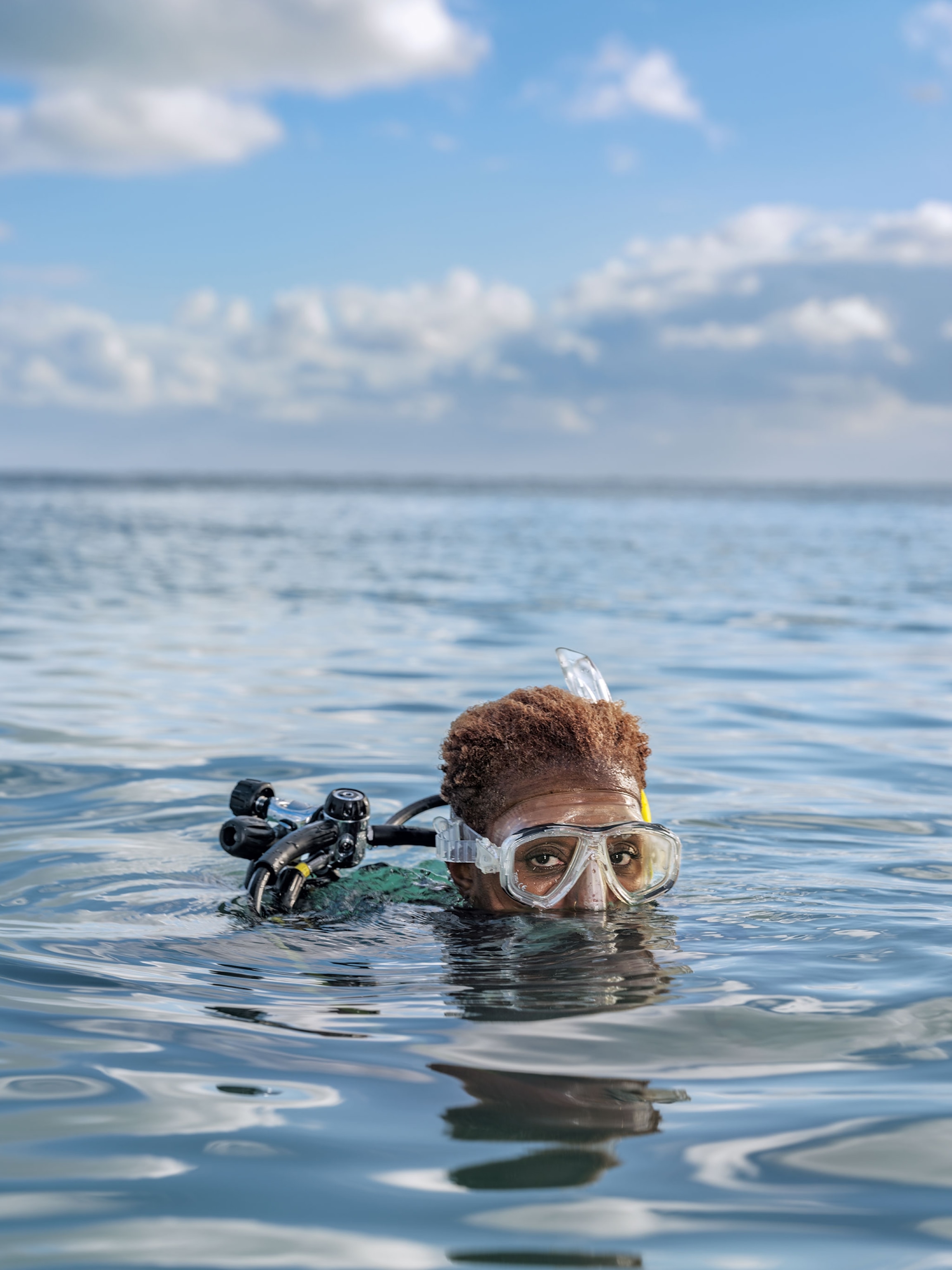 A woman in full scuba gear pops her head out of water looking at the camera.