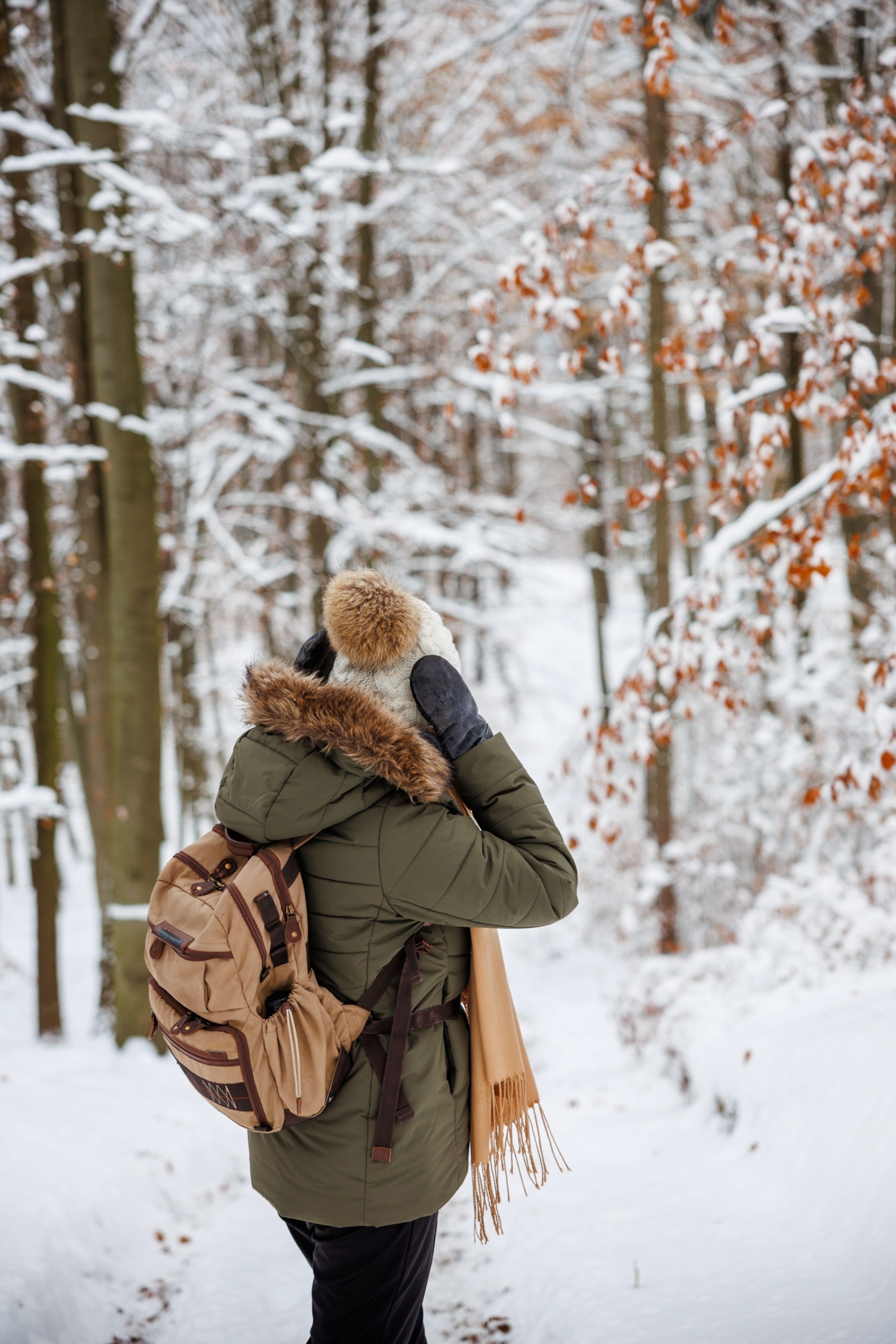A woman wearing an olive green down jacket and tan backpack is seen from behind while standing in snow-covered woodlands. She is adjusting her white knitted hat with mitten covered hands.