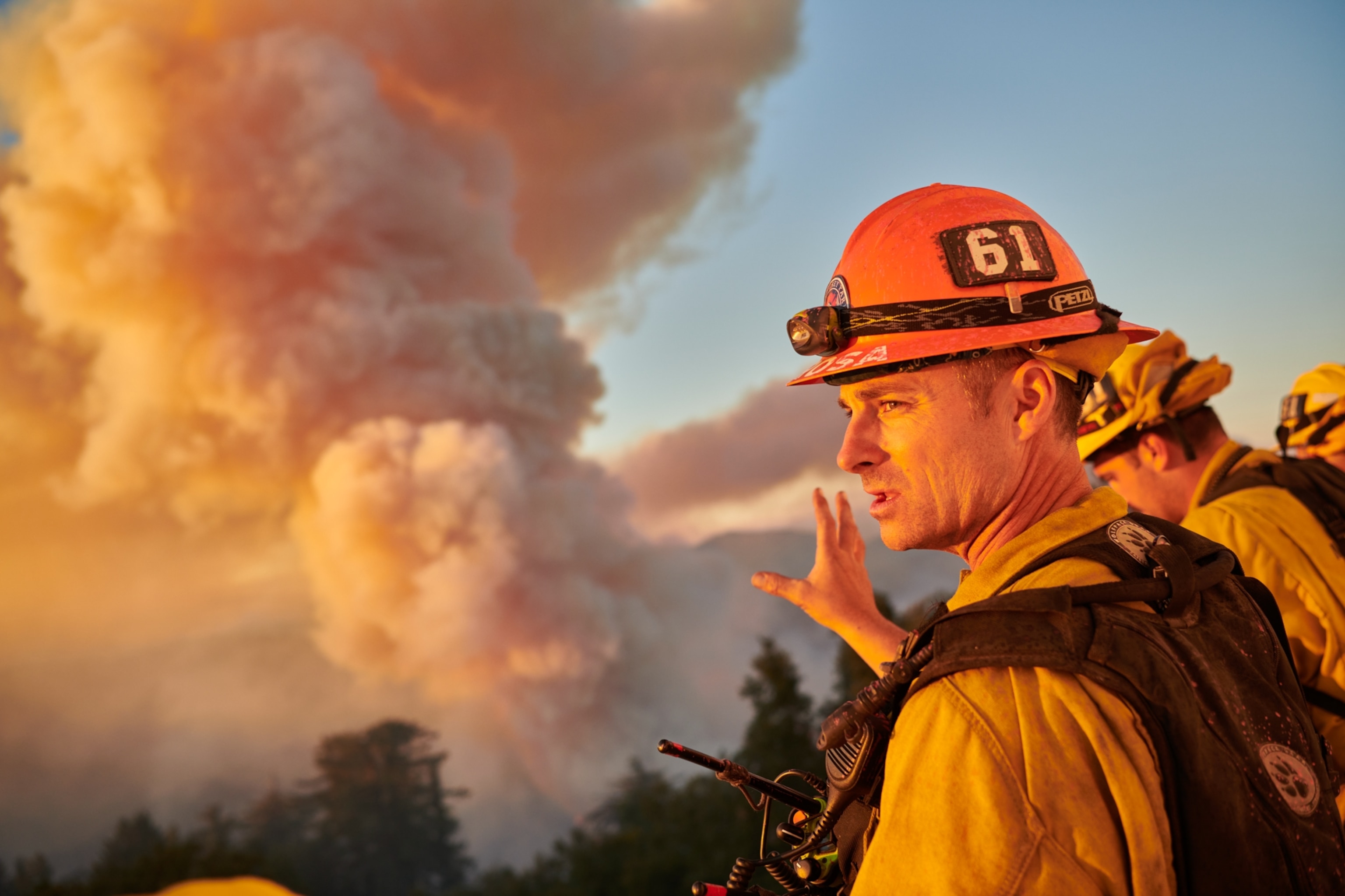 Firefighters atop a high point overlooking a huge plume of smoke rising from the forest below.