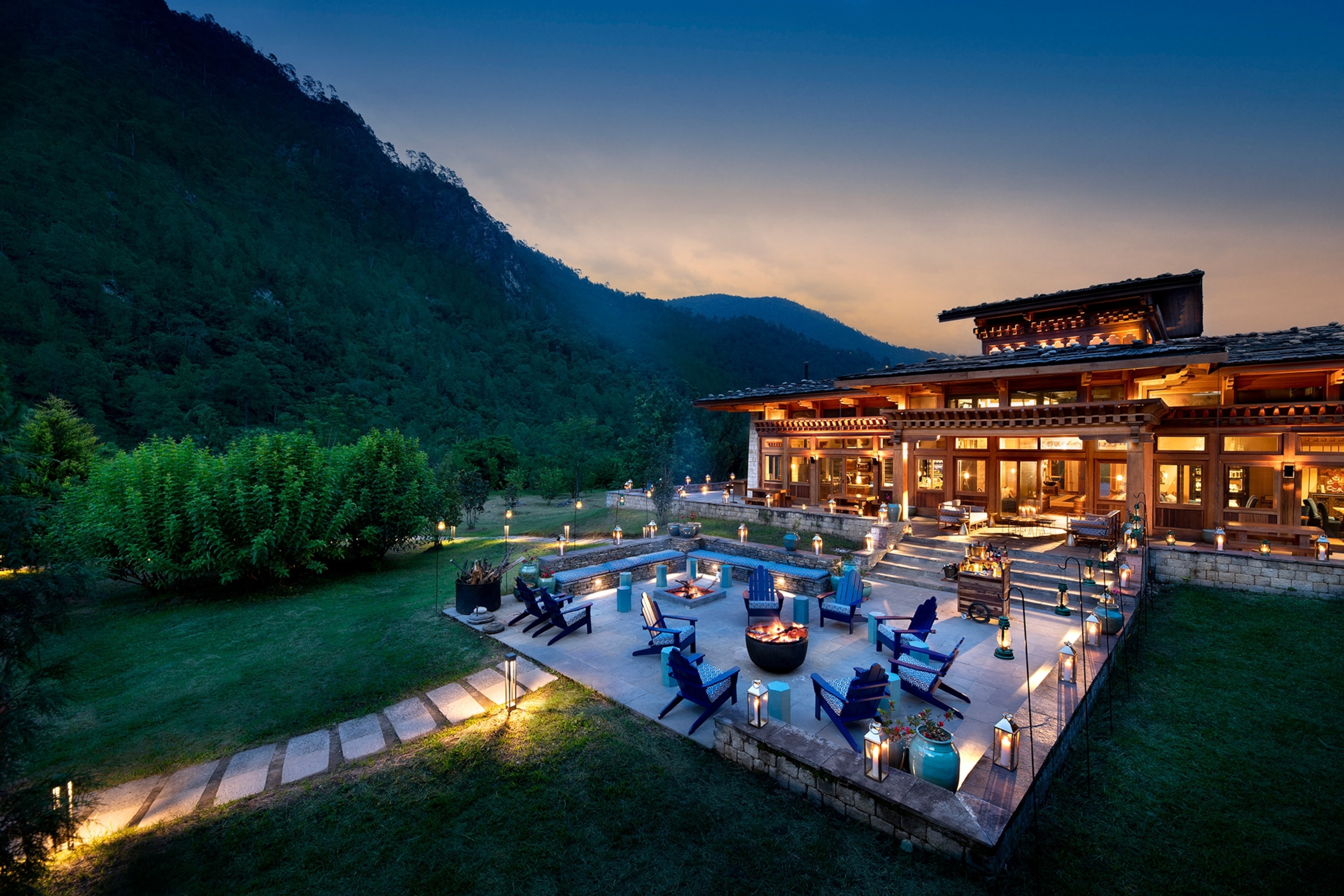 An evening view of the andBeyond Punakha River Lodge and fire pit nestled in the Himalayan foothills. Warm lights illuminate the inside.