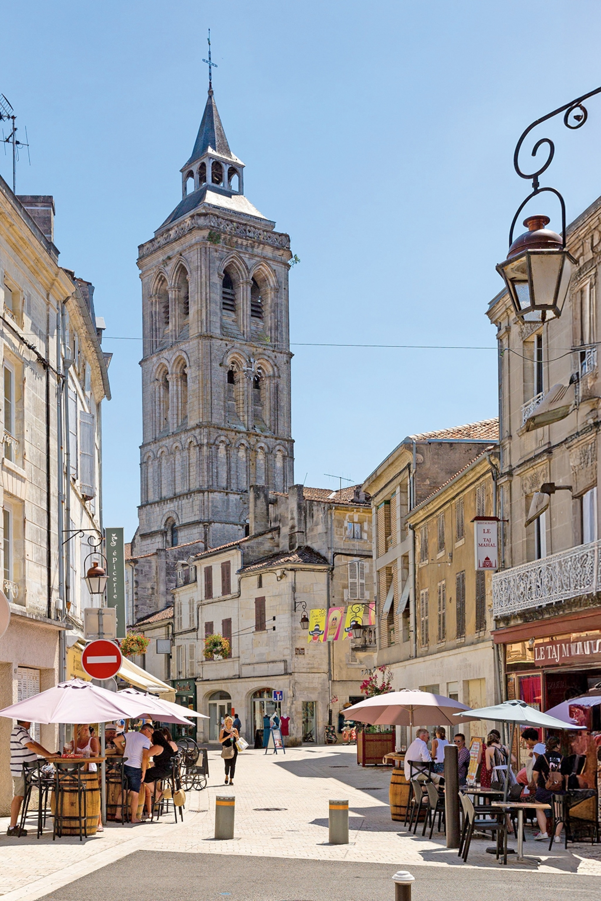 A view down the pedestrian street of a low-roofed, traditional French town with a church tower in the background.