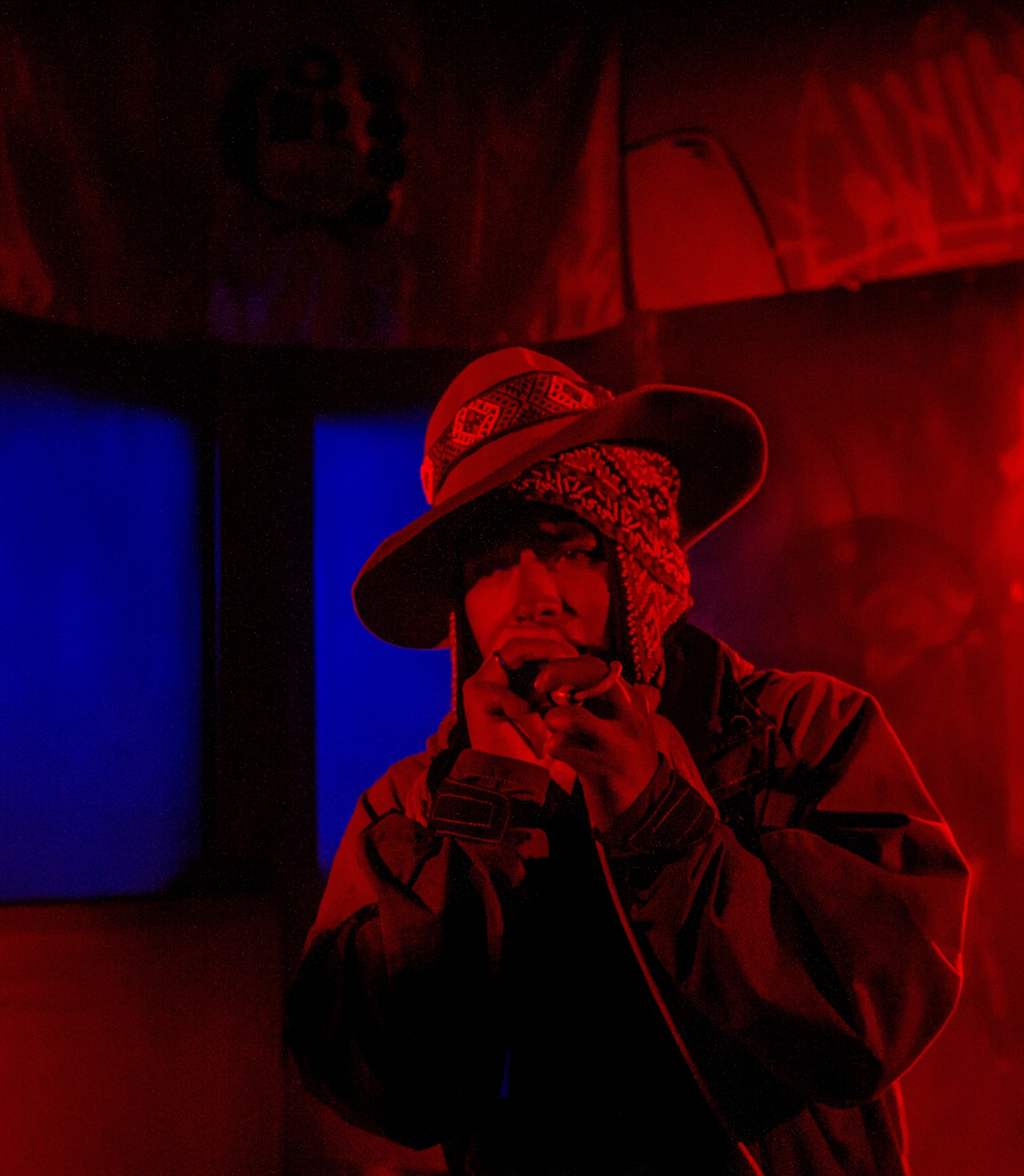Javier Cruz is holding a microphone singing. He is wearing a hat and a jacket. The lighting is red with blue windows.